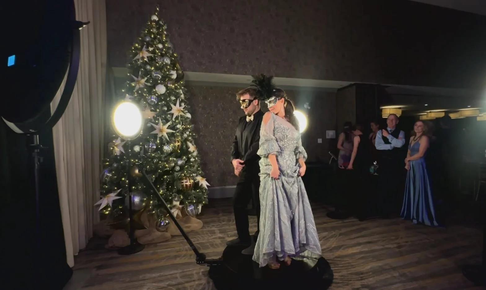 People in elegant costumes wearing masquerade masks dancing near a decorated Christmas tree at an indoor party.