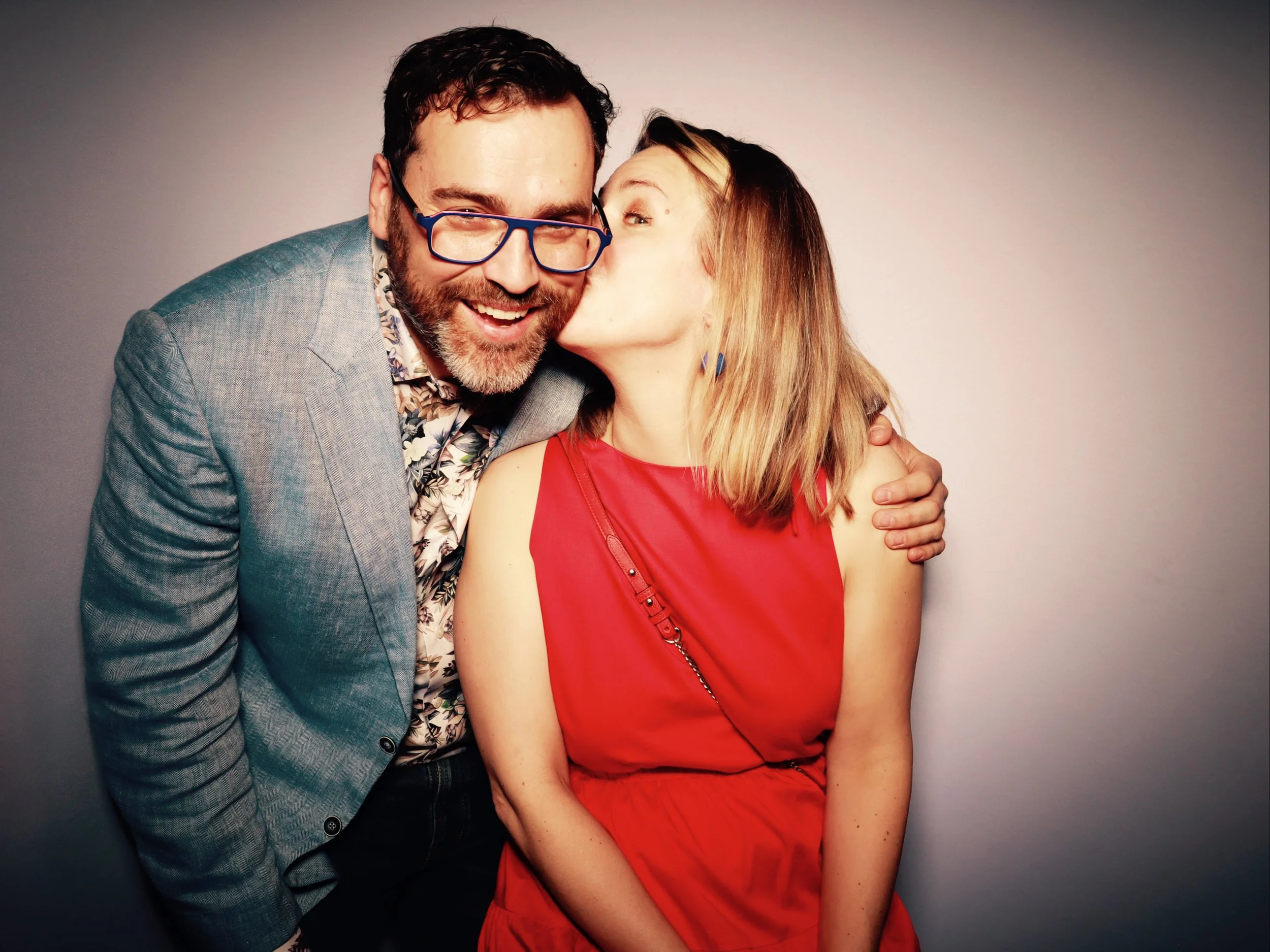 A man with glasses and a beard in a light blazer is giving a woman in a red dress a kiss on the cheek, both smiling and standing close together against a plain background.