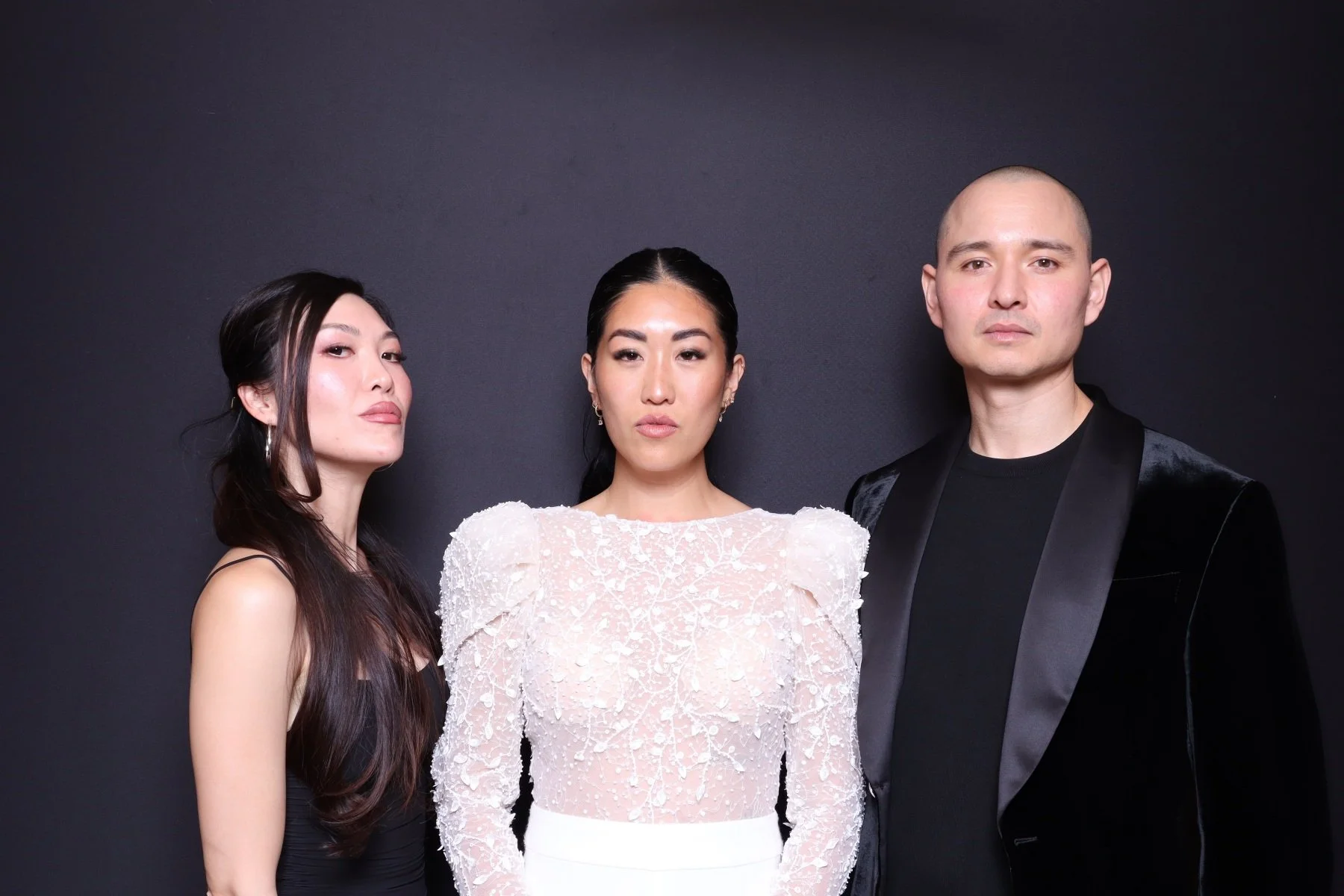 Three fashionably dressed individuals posing against a dark background, with the two women on either side and a man in the center.