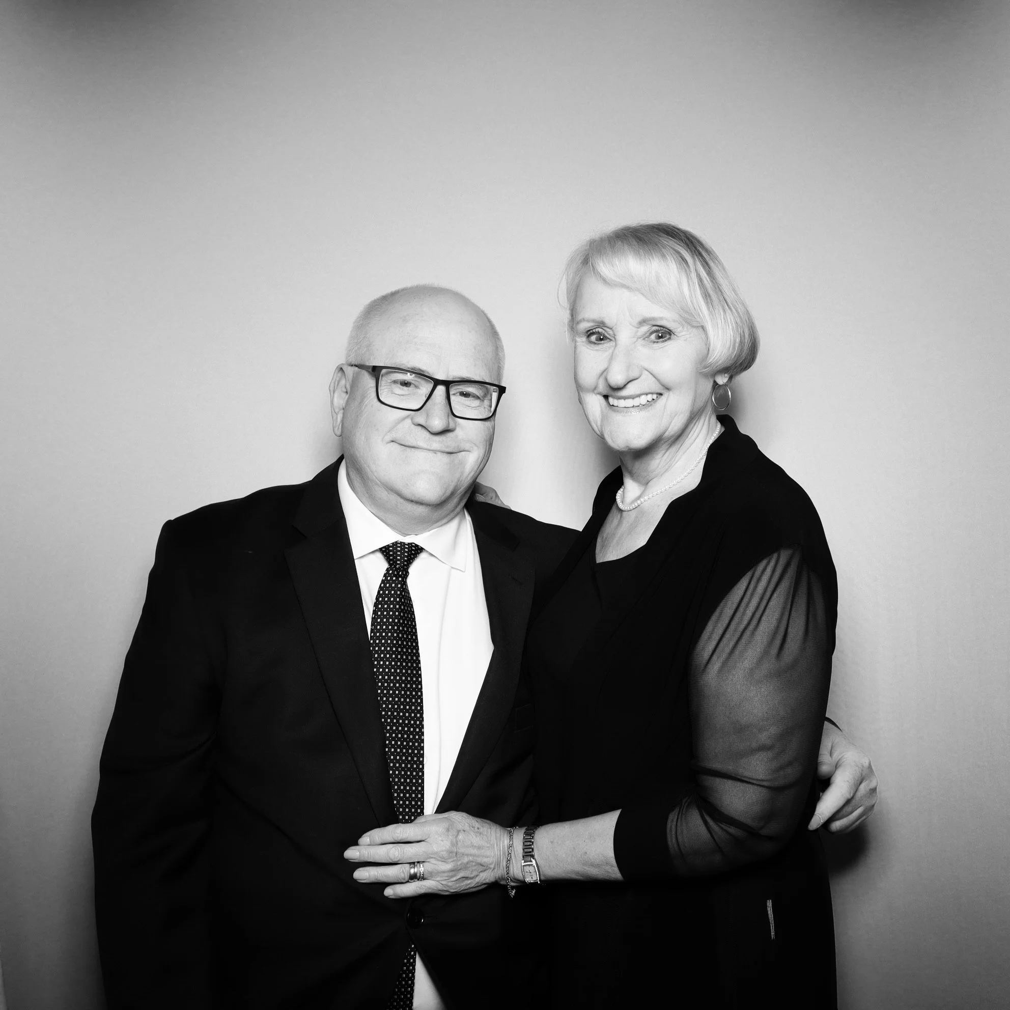 Black and white photo of a smiling older man and woman standing close together, the man wearing glasses and a suit, the woman wearing a black dress with sheer sleeves, against a plain background.