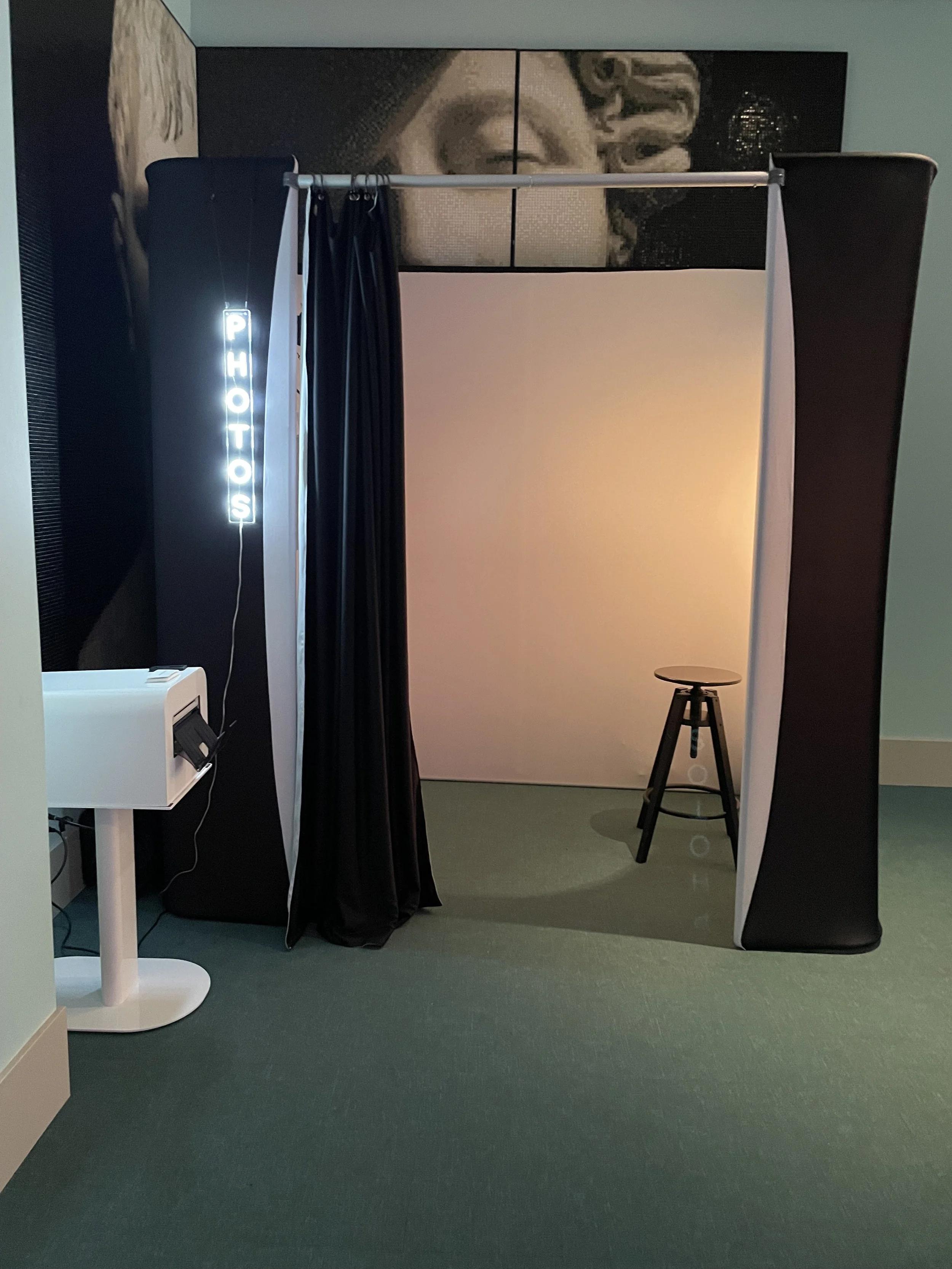 Photo booth with black curtains, a stool, and a white backdrop, with a neon sign reading 'PHOTOS' hanging on the left side.