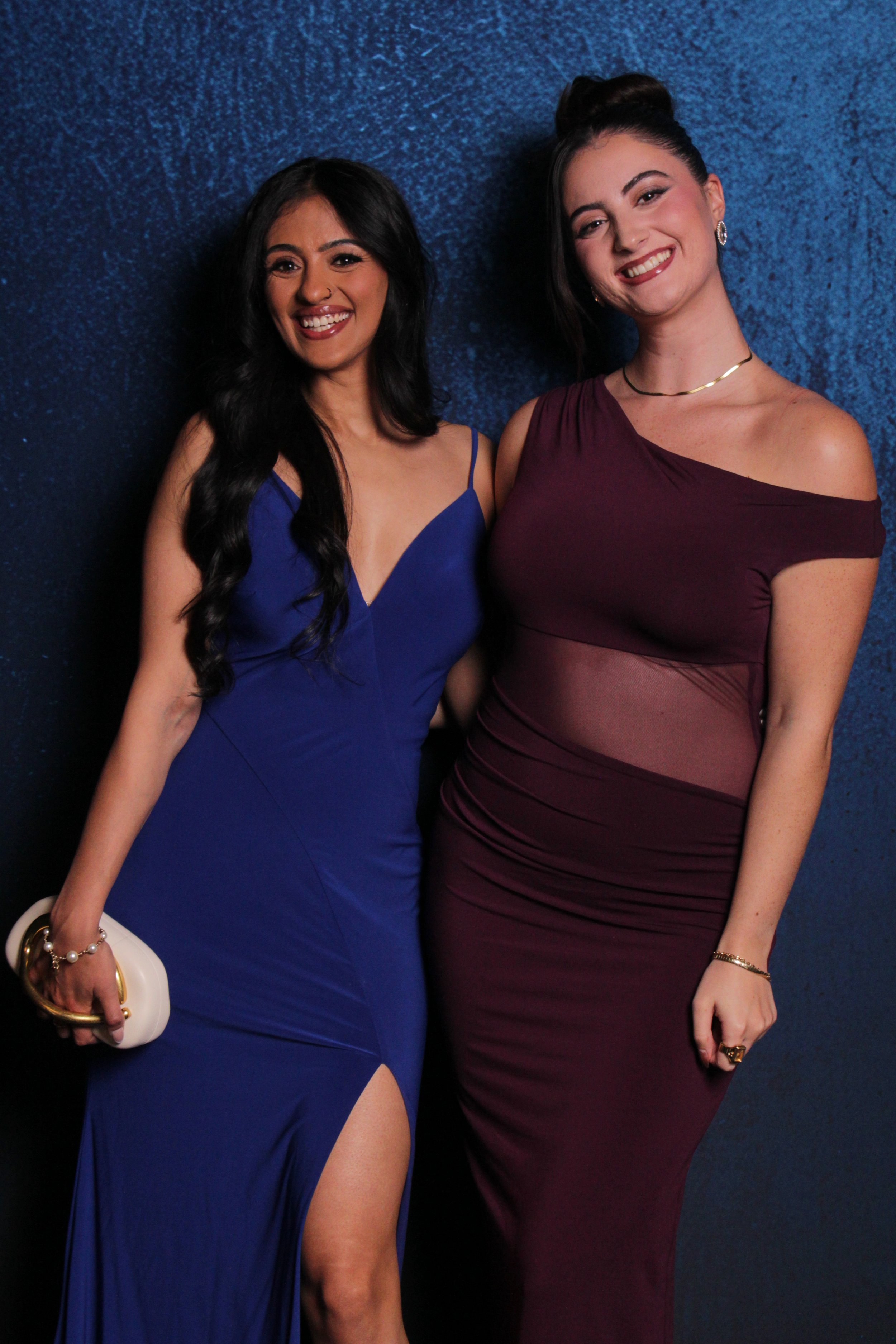 Two women dressed in elegant gowns standing together and smiling against a blue wall. The woman on the left wears a blue dress with a thigh-high slit, holding a clutch, and has long wavy black hair. The woman on the right wears a burgundy dress with 