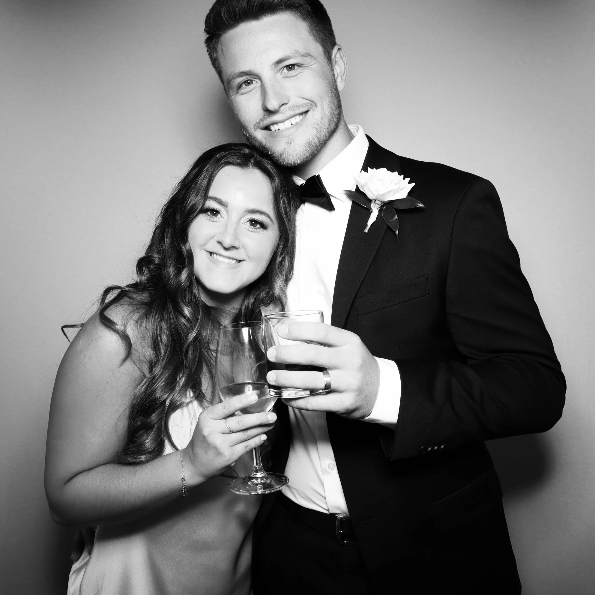 A smiling couple in formal attire, holding glasses of wine, posing together against a plain background.