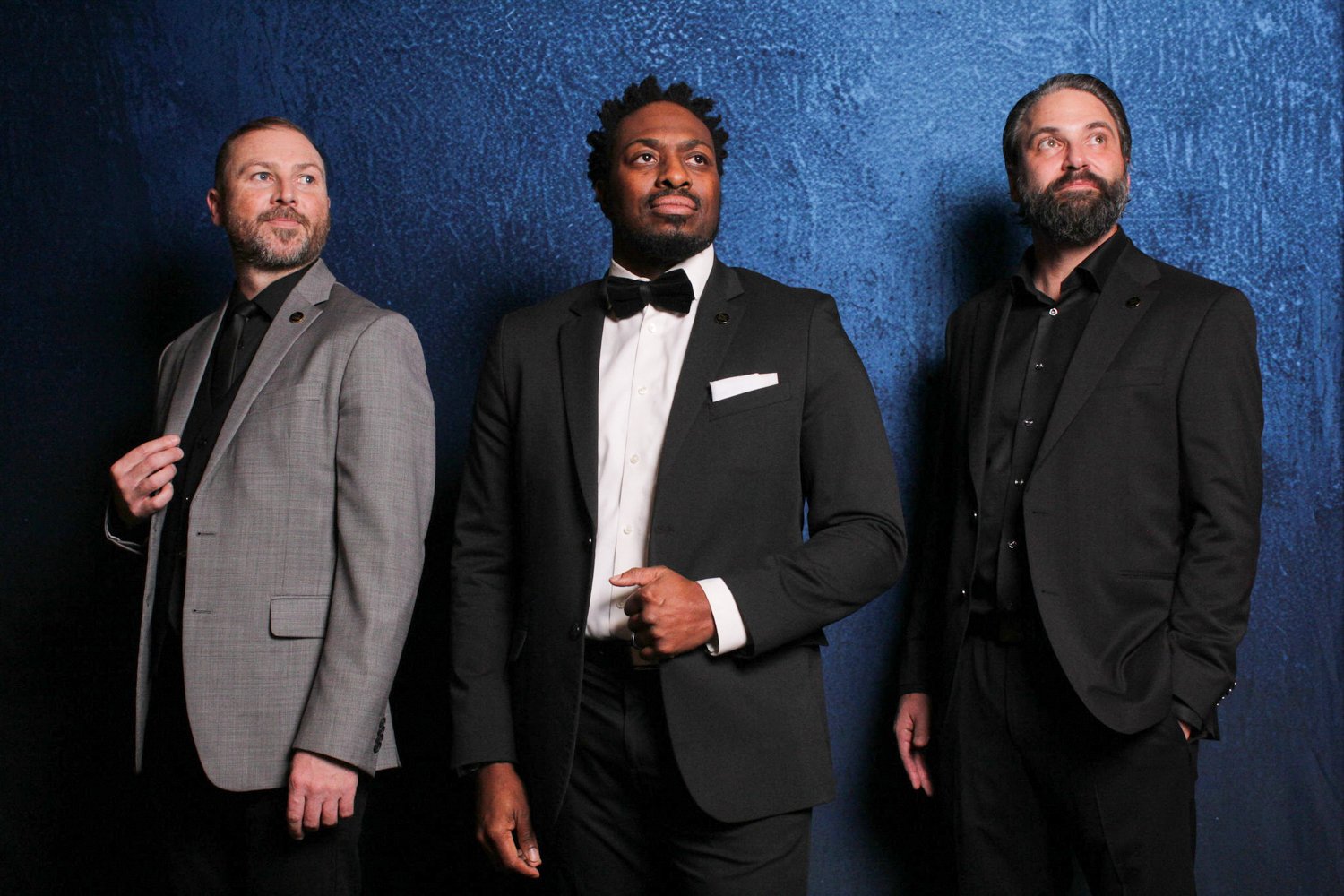 Three men in formal suits standing against a blue textured wall, with the man on the left in a gray plaid suit, the man in the middle in a black tuxedo with a bow tie, and the man on the right in a black suit.