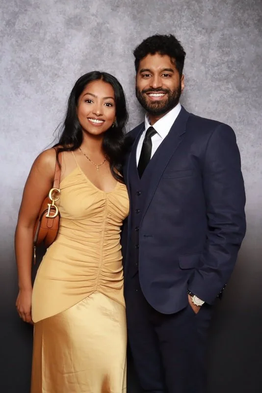 A smiling woman in a gold dress and a man in a dark navy suit standing together against a gray textured background.