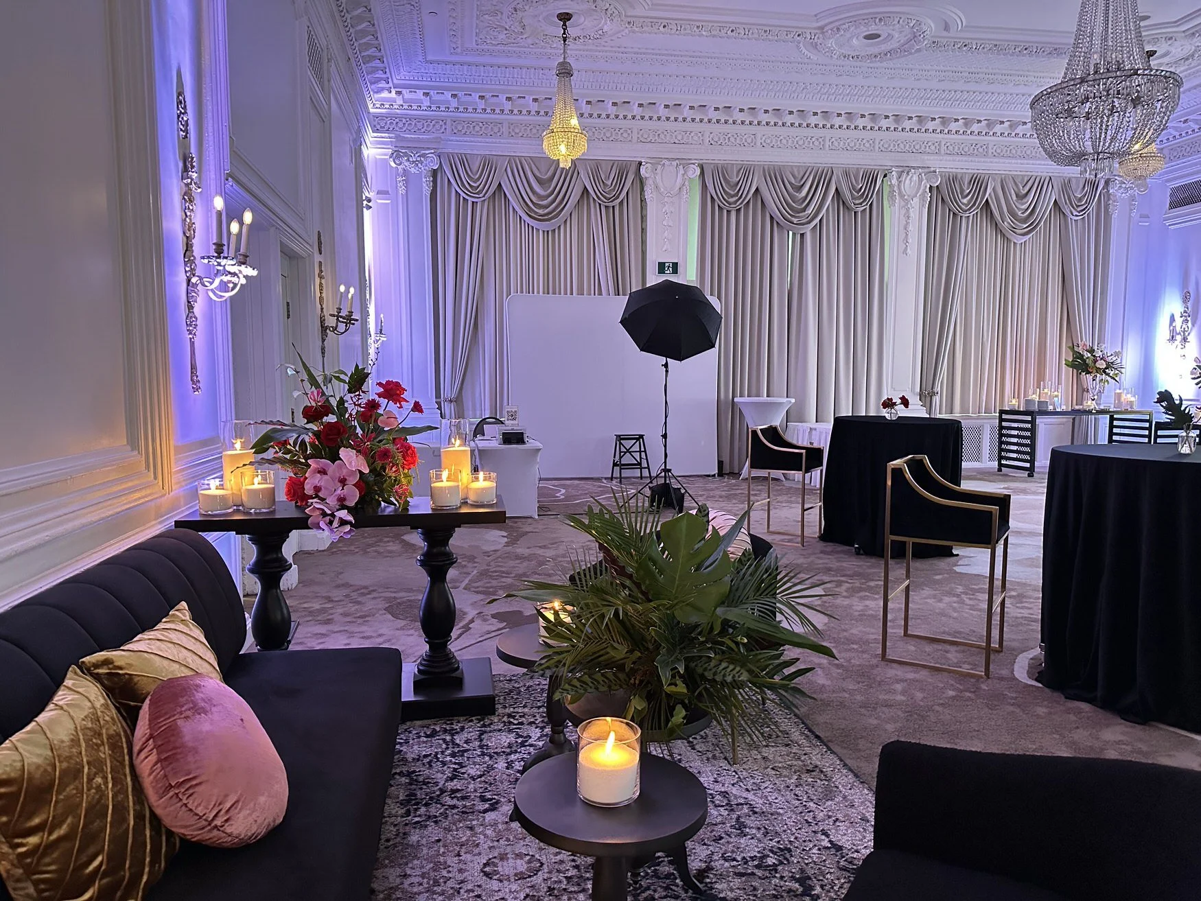Elegant event room with ornate ceiling and chandeliers, decorated with beige drapes, floral arrangements, candles, and black tables, ready for a celebration or ceremony.