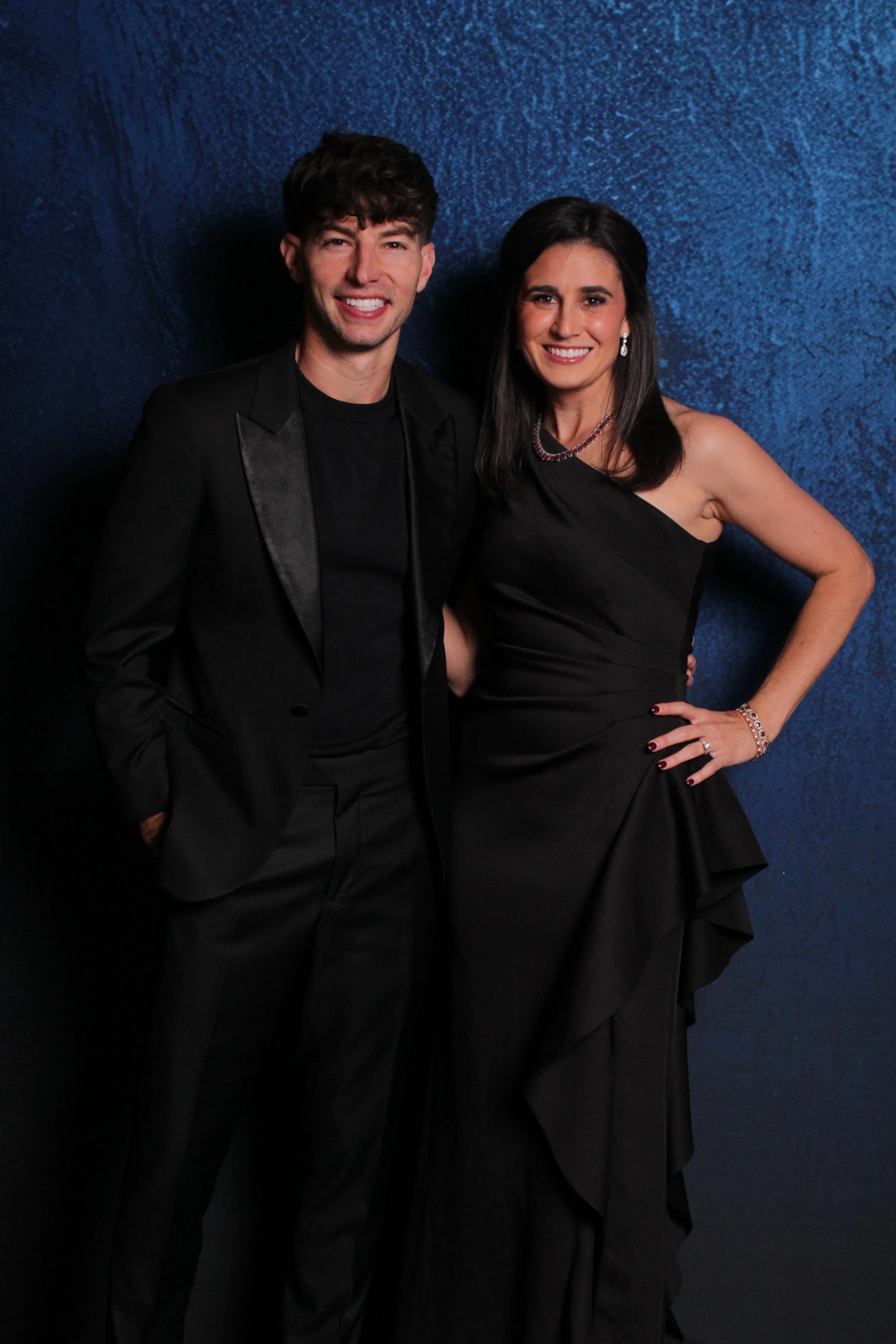 A man and woman dressed formally in black, standing close together against a textured blue wall, smiling at the camera.