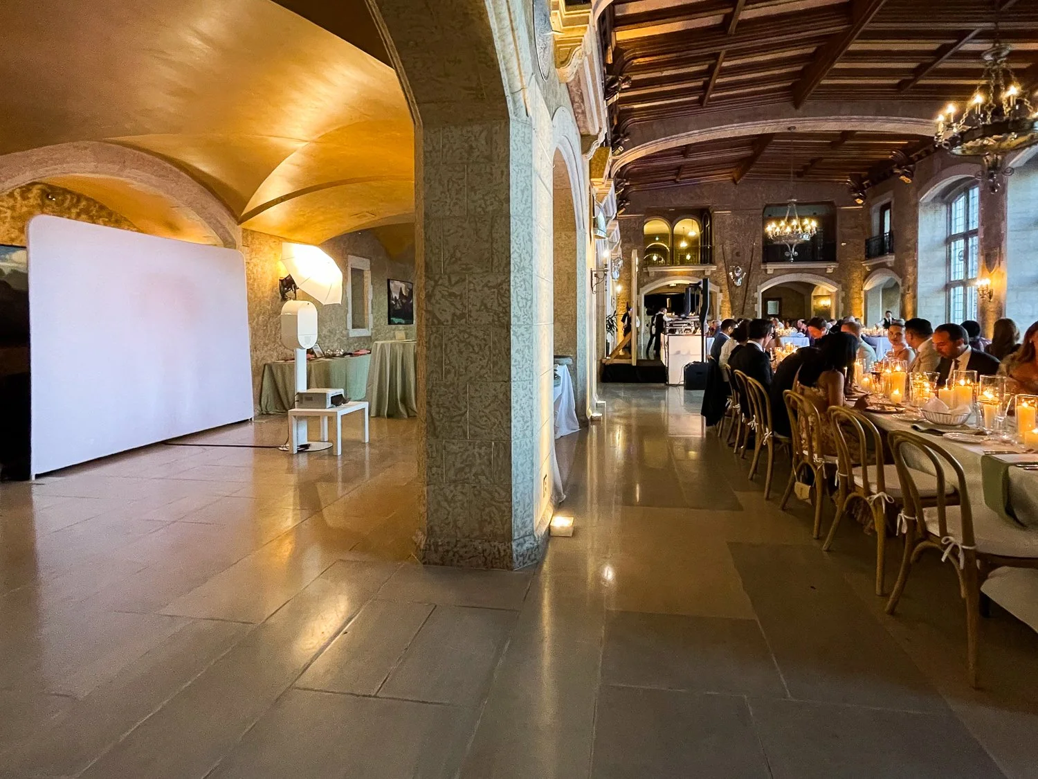 Elegant banquet hall with high wooden ceiling, stone walls, large windows, and candles on tables. Guests are seated at a long dining table with white chair covers, enjoying a formal dinner.