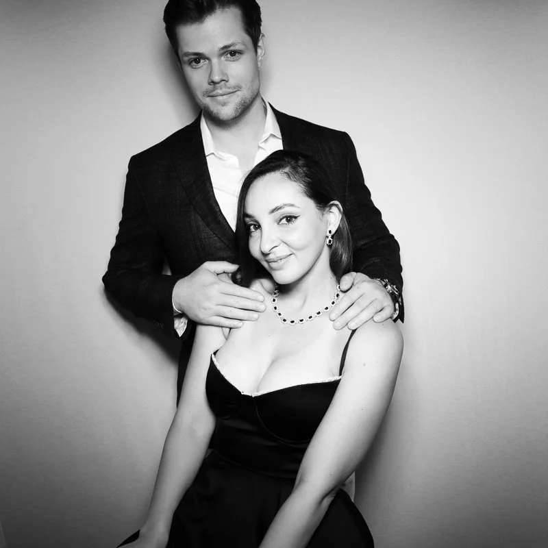 A black and white photo of a man and woman posing together. The man is standing behind the woman with his hand on her shoulder, both are dressed formally with the woman wearing a dress and jewelry, and the man in a suit.