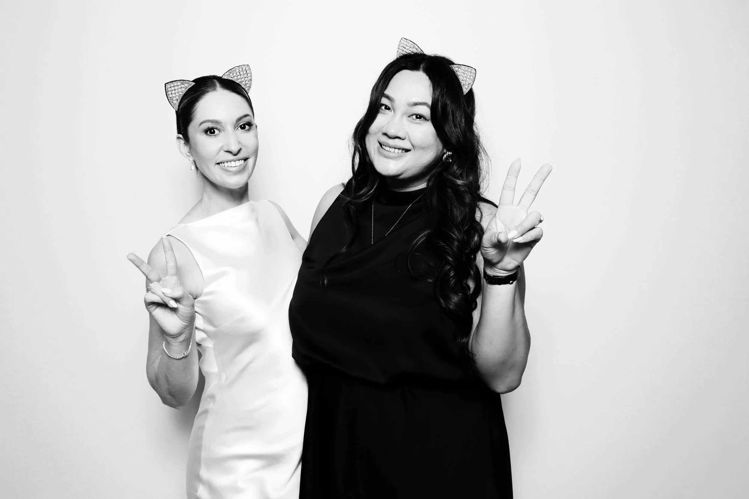 Two women smiling, wearing cat ear headbands, making peace signs, black and white photo.