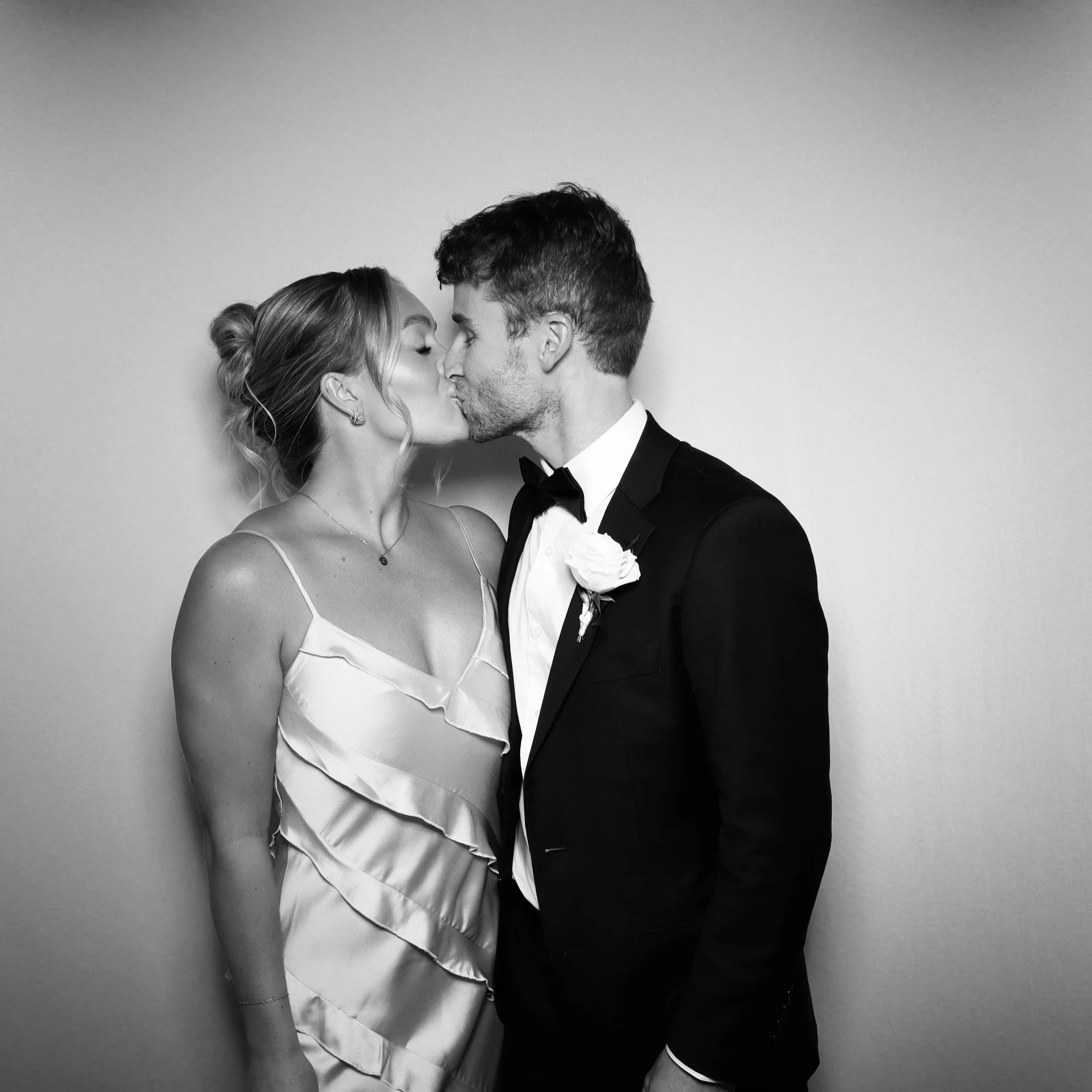 A black and white photo of a bride and groom sharing a kiss, dressed in wedding attire, against a plain background.