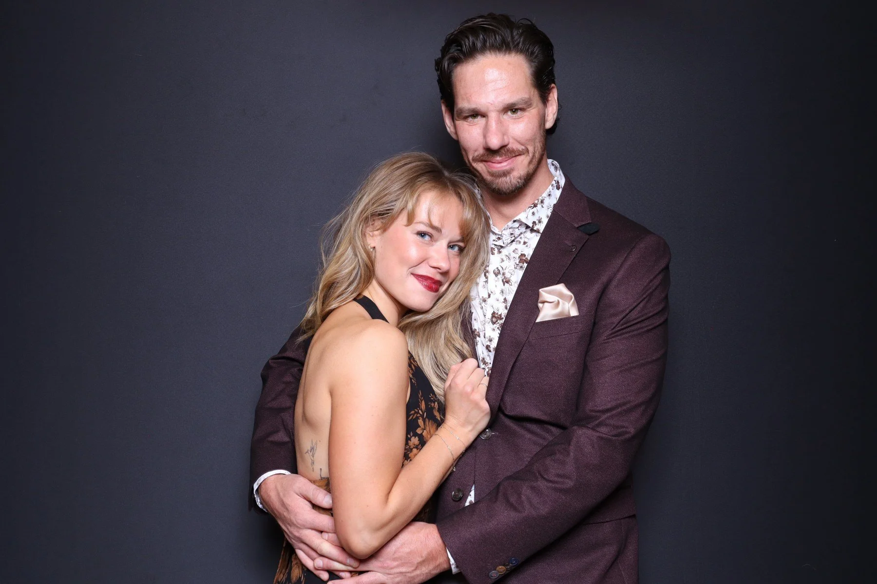 A man and a woman standing close together, smiling, against a plain dark background. The woman has blonde hair, and the man has dark hair and a beard, wearing a maroon suit with a patterned shirt and pocket square.