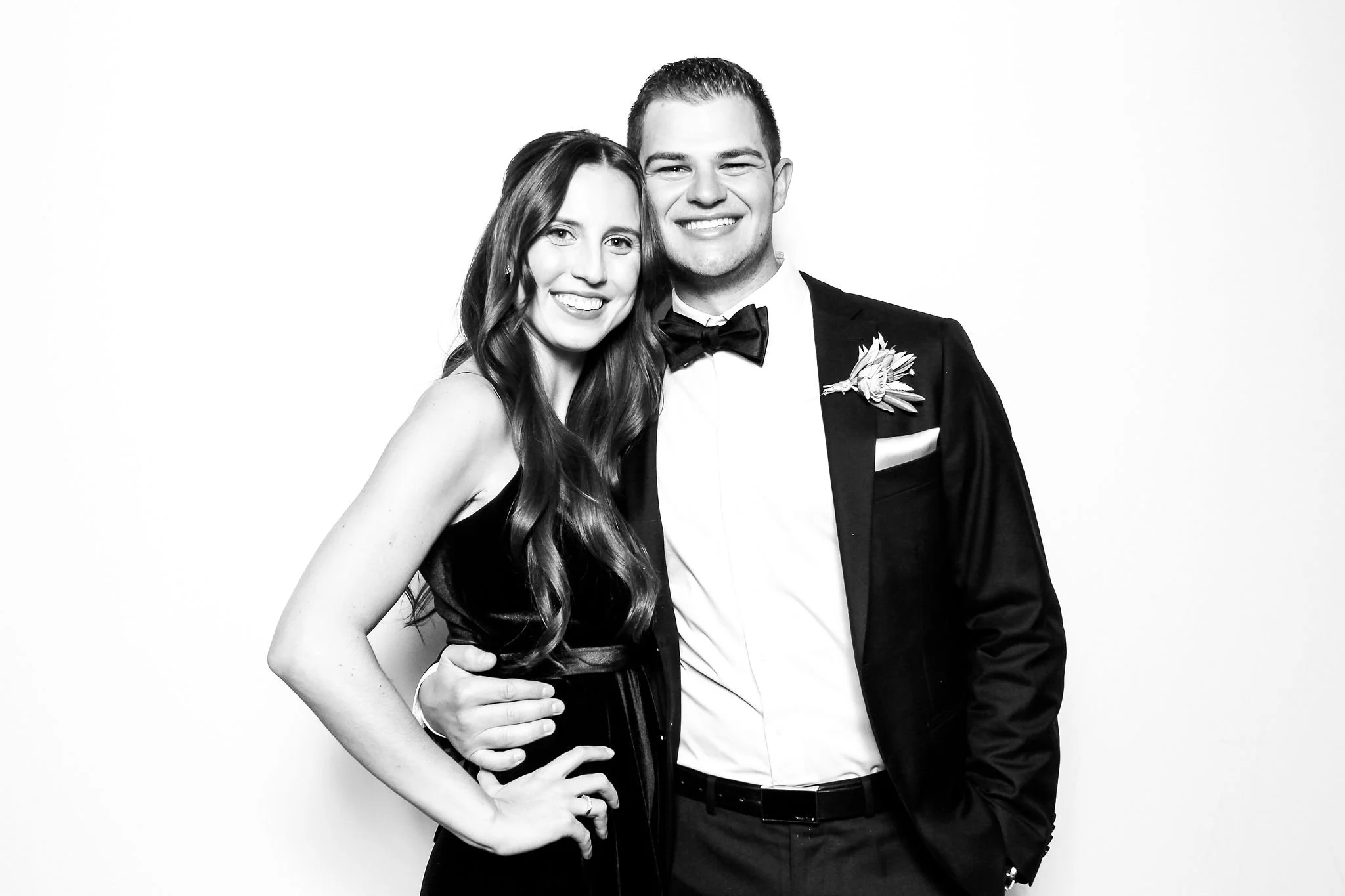 A young couple dressed in formal attire, with the woman in a black dress and the man in a tuxedo, smiling and posing together.