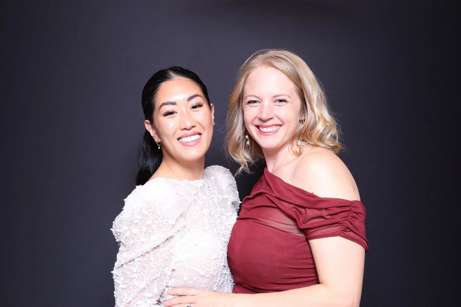 Two women smiling and standing close together against a dark background, one with black hair in a white dress, the other with blonde hair in a red dress.