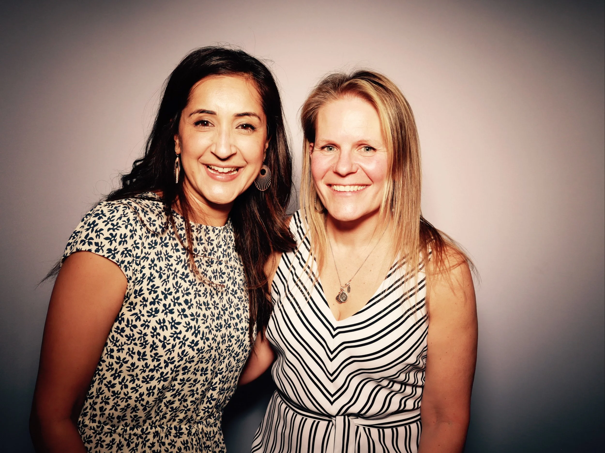 Two women smiling and posing together in front of a neutral background, one with dark hair and the other with blonde hair, wearing striped and floral tops.