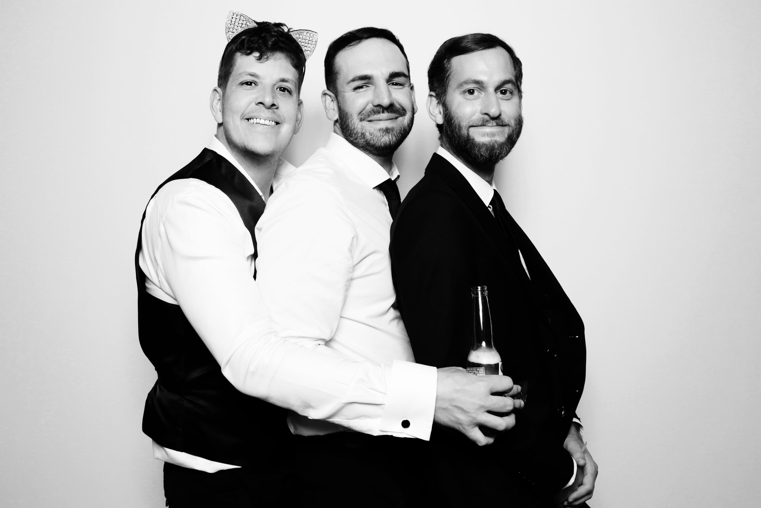 Three men standing in a row, dressed in formal attire, posing against a plain background, with one holding a bottle, in black and white photo.