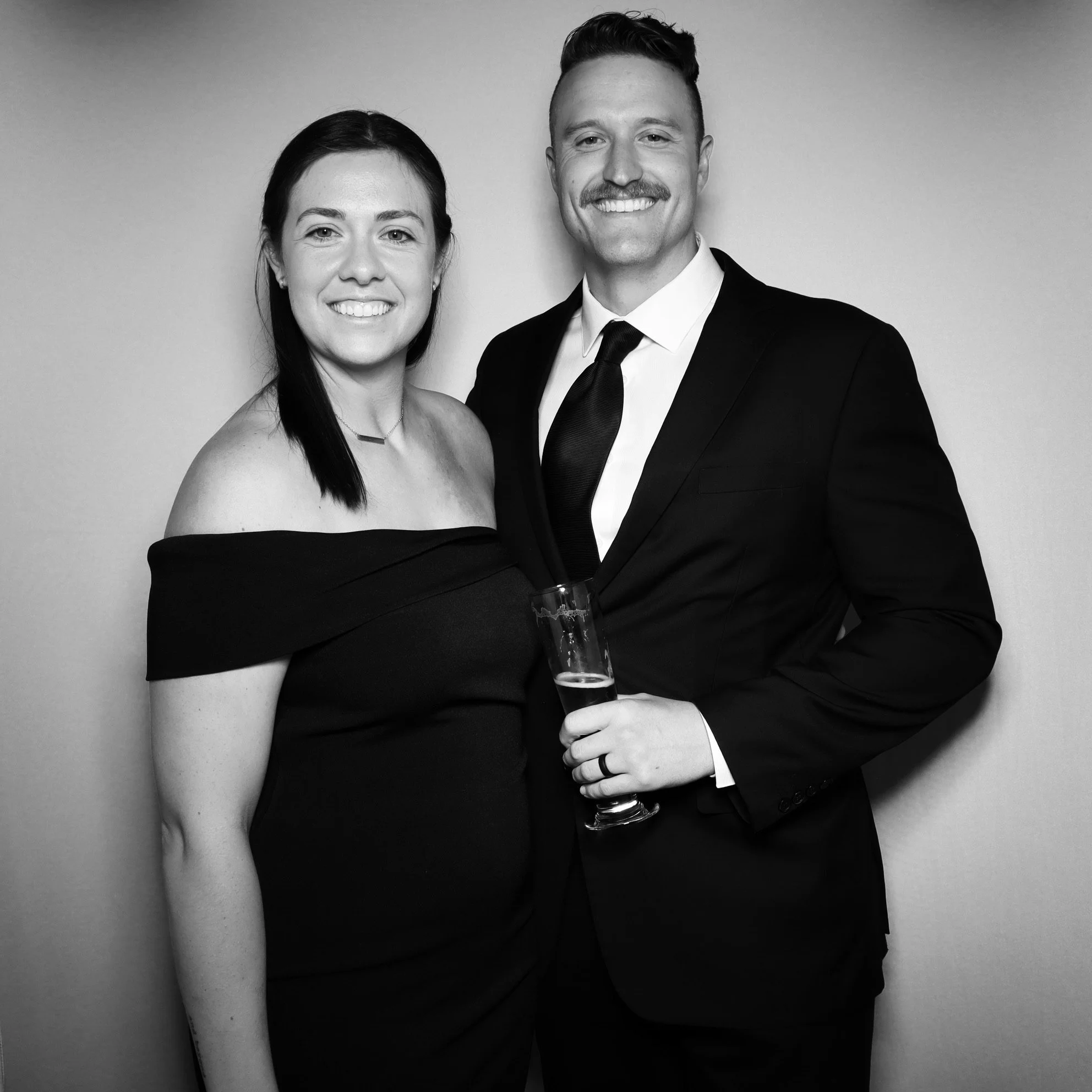 A smiling woman in an off-the-shoulder black dress standing next to a smiling man in a black suit and tie, holding a drink, against a plain wall.