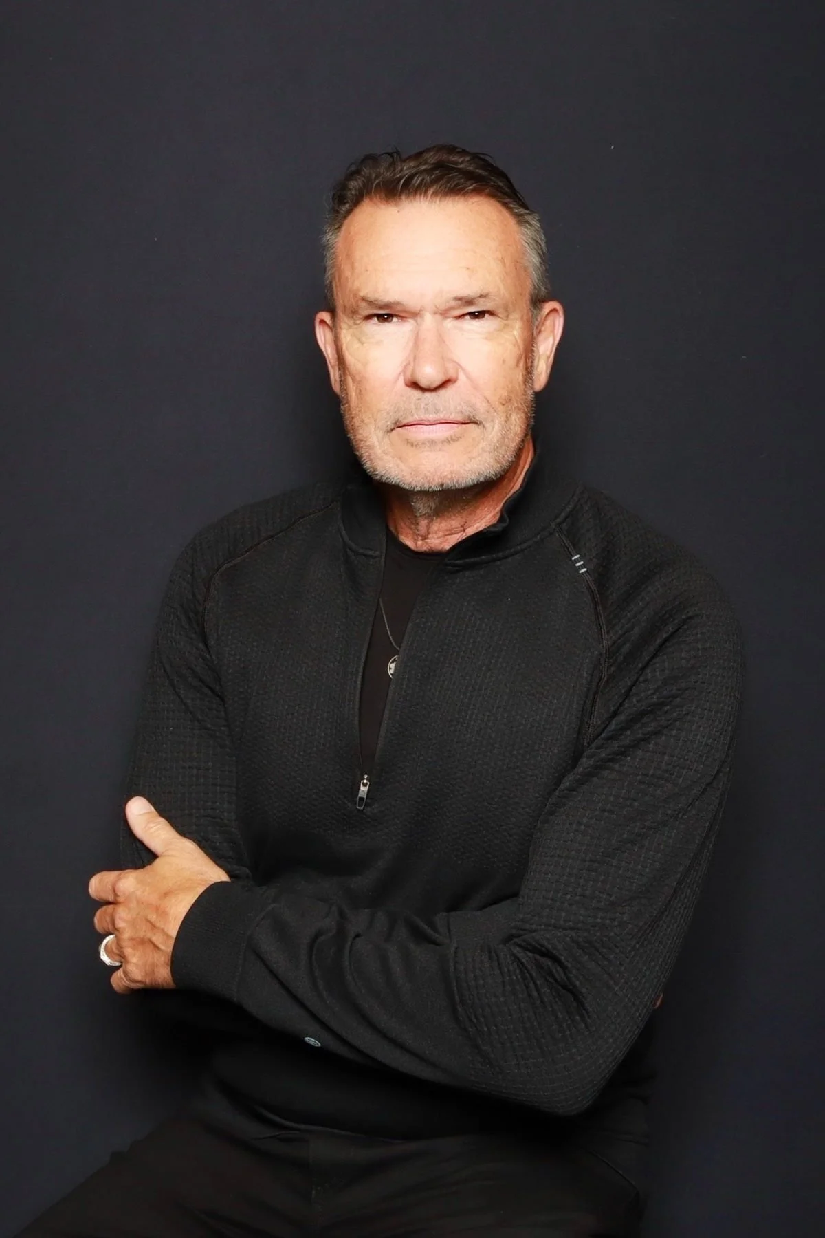 A middle-aged man with gray hair and a beard, wearing a black zip-up jacket, sitting against a dark background with arms crossed, looking directly at the camera.