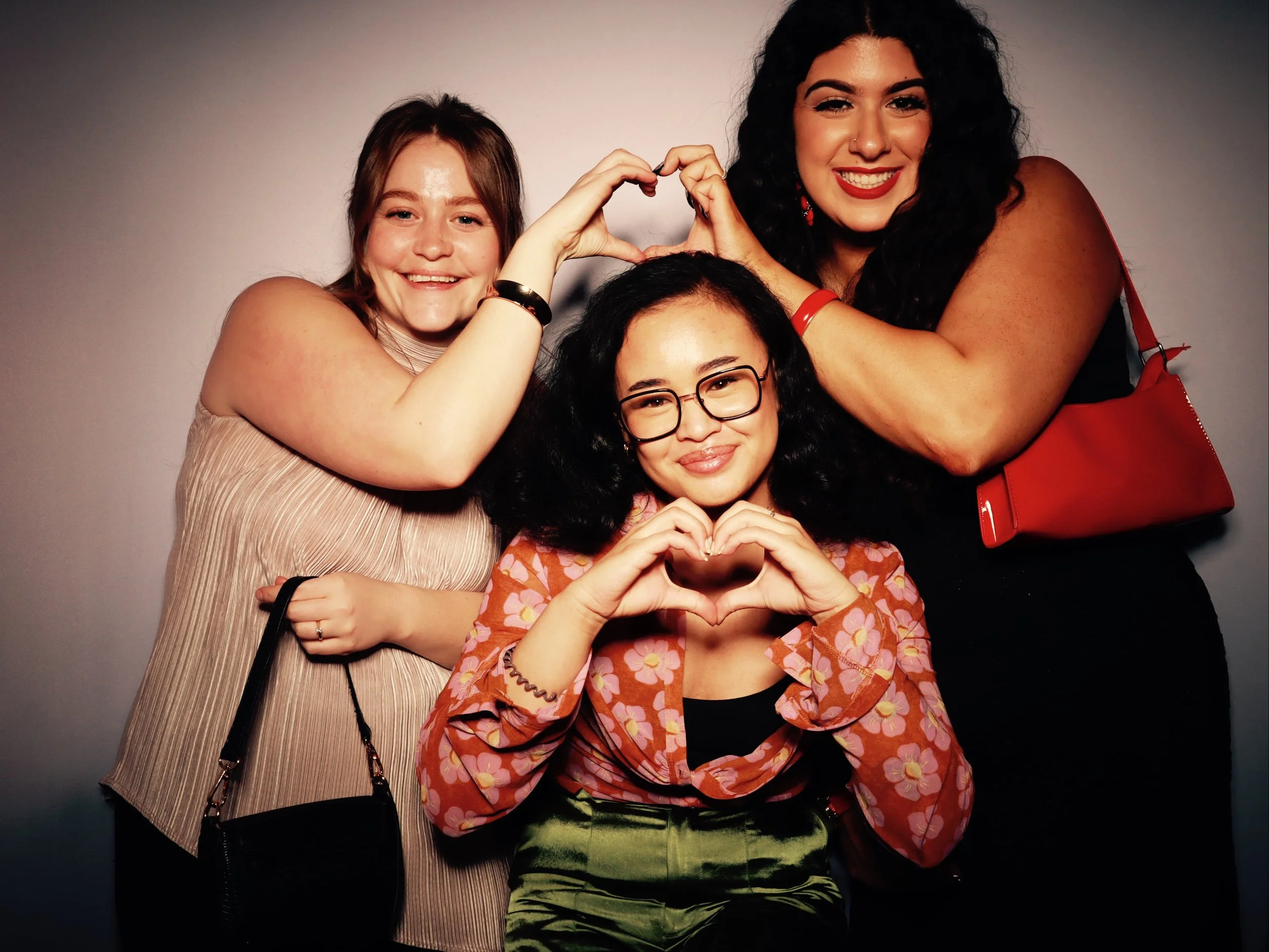 Three smiling women making heart shapes with their hands, with one woman in the center and two women behind her, all against a plain background.