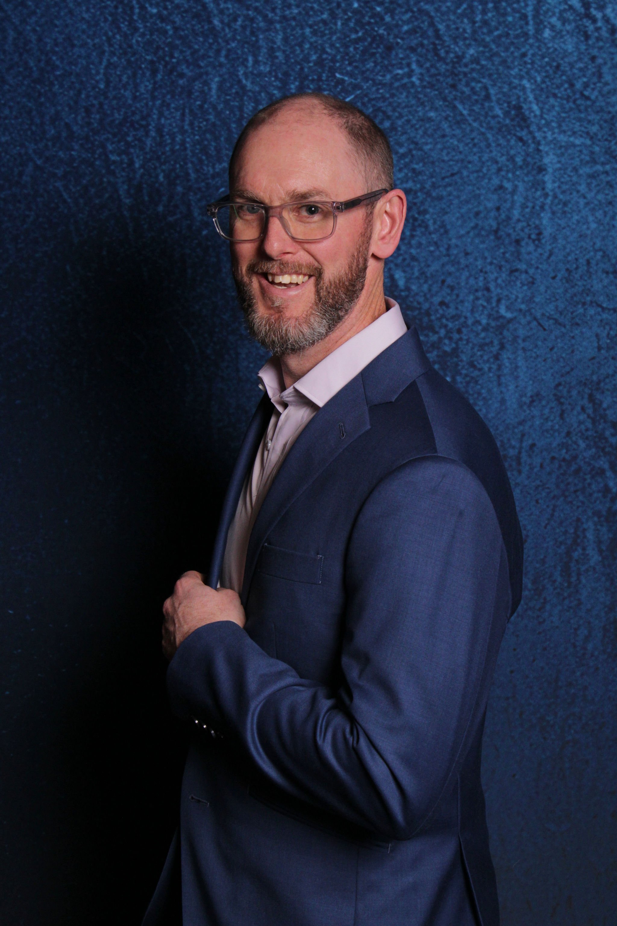 A man in a blue suit and glasses posing against a textured blue background.