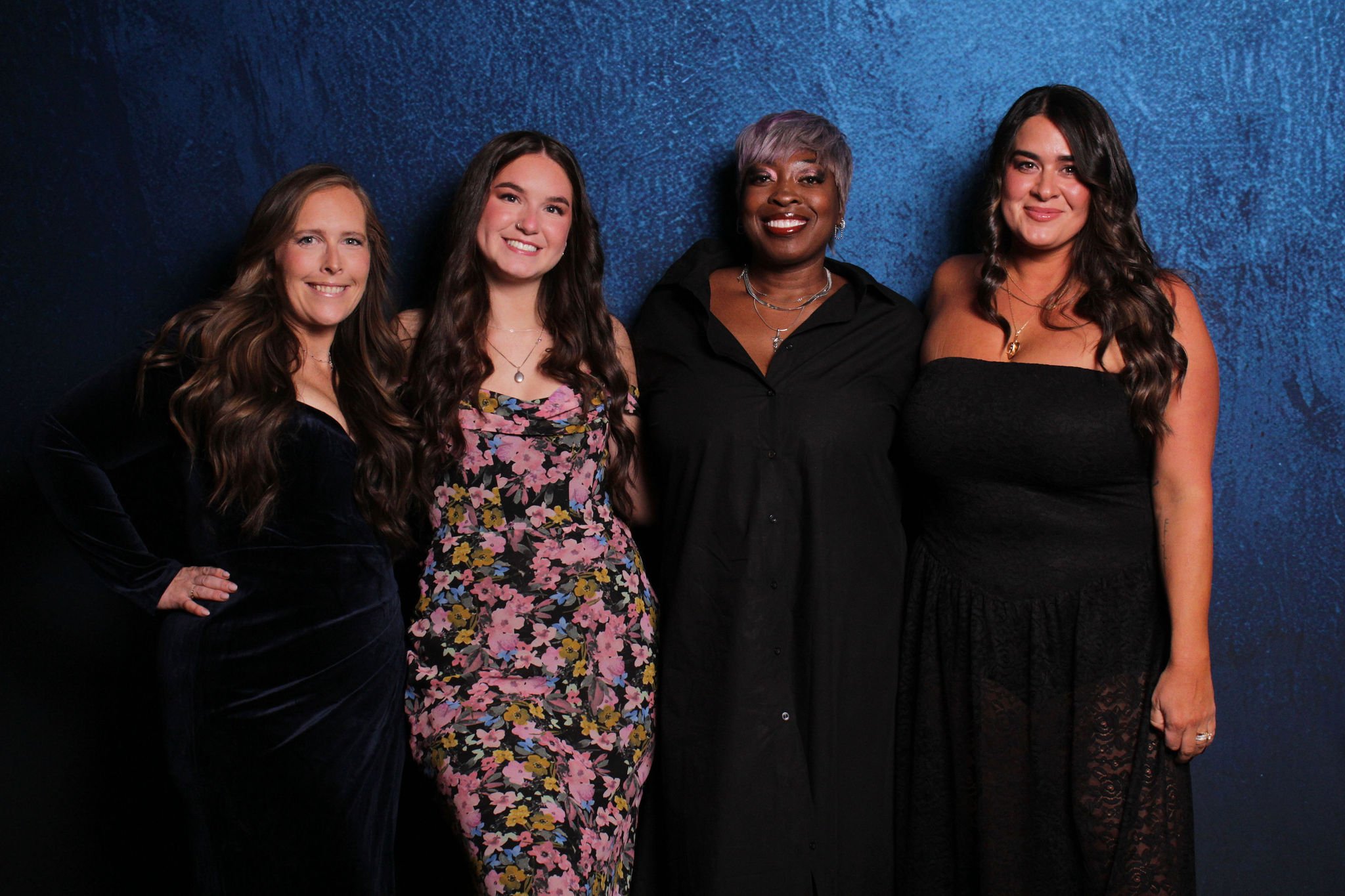 Four women standing side by side against a blue textured background, smiling at the camera.
