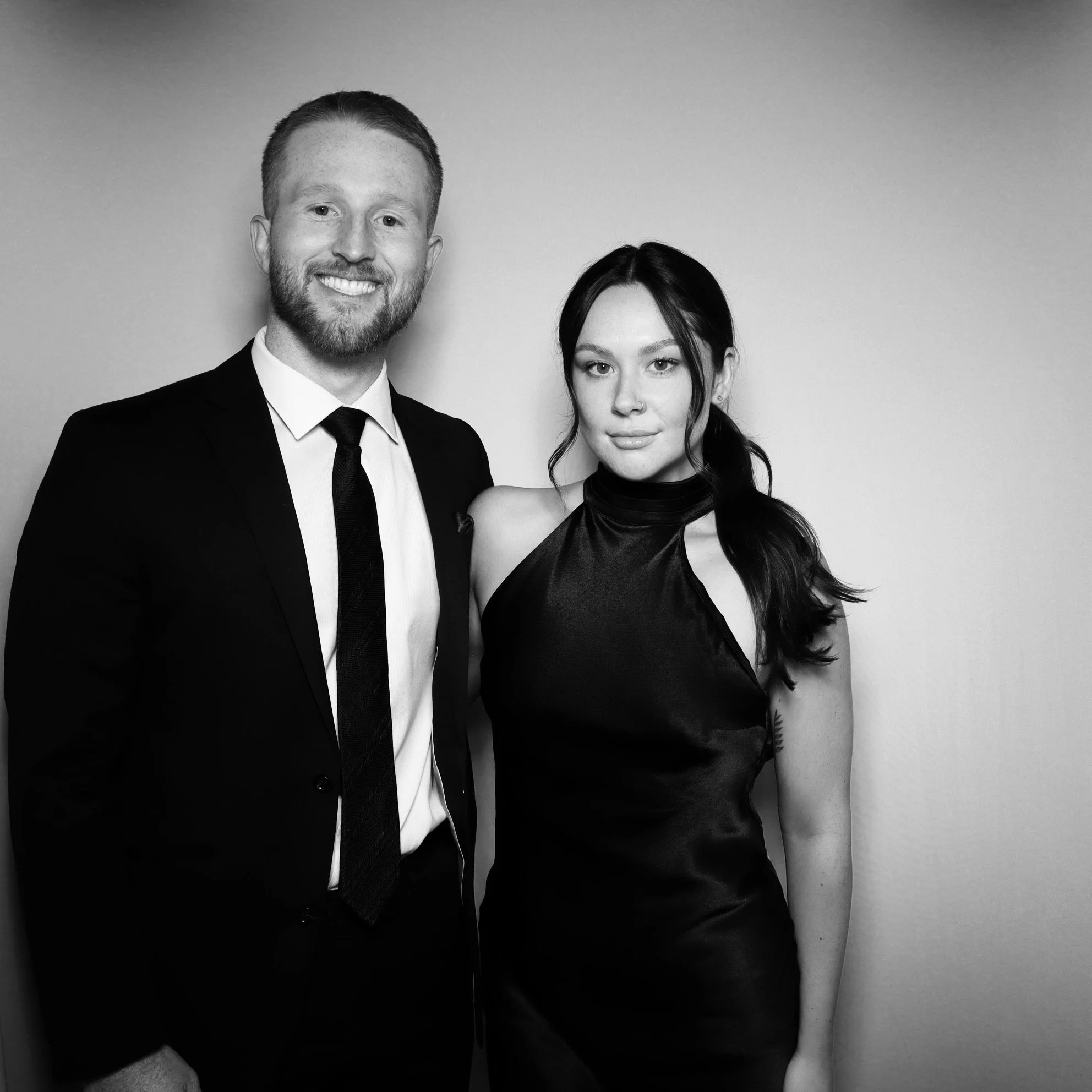 A smiling man in a suit and a woman in a shiny dress standing together in front of a plain wall.