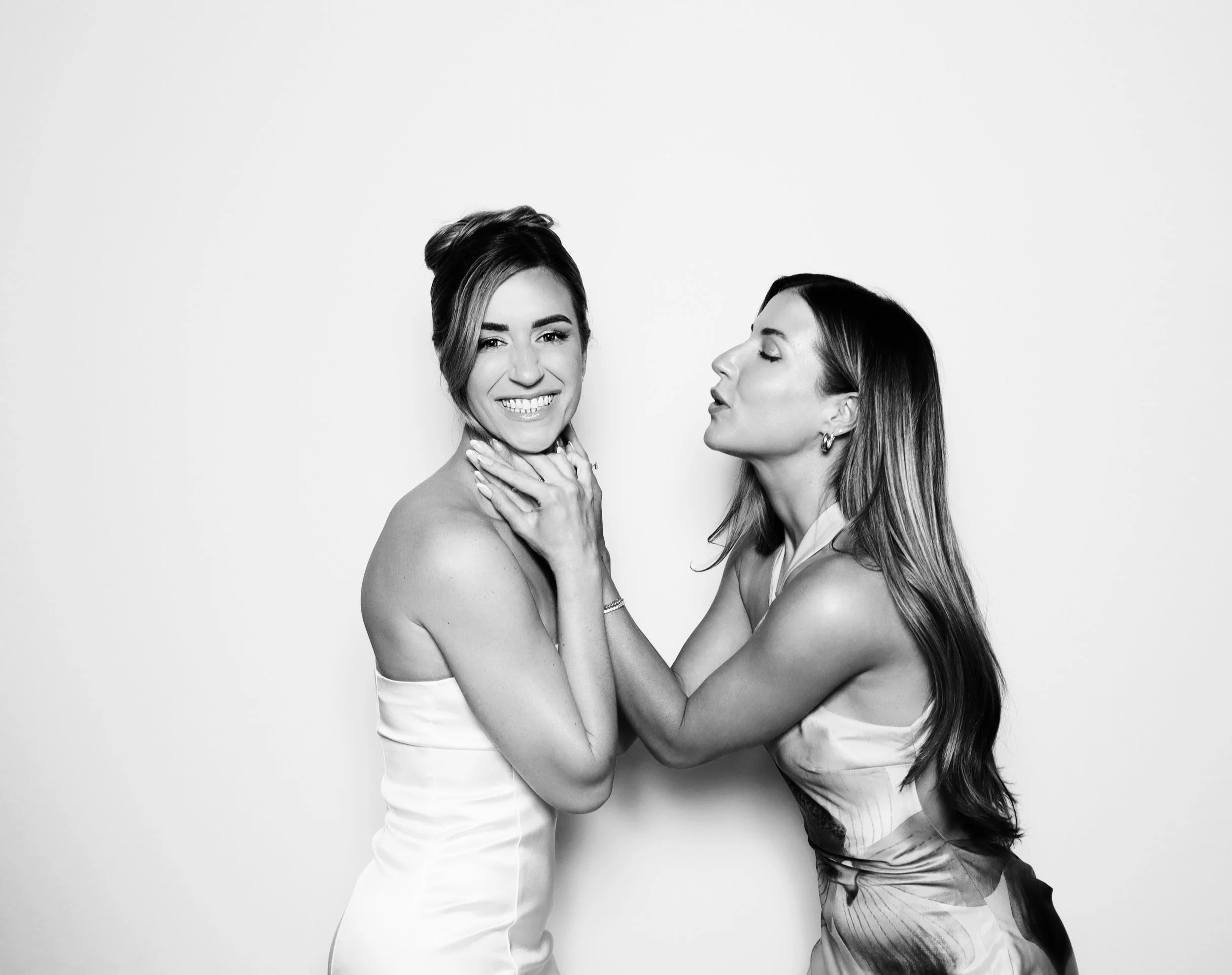 Two women in elegant dresses facing each other; one woman smiling and the other woman appearing to whisper or kiss her cheek, against a plain background.