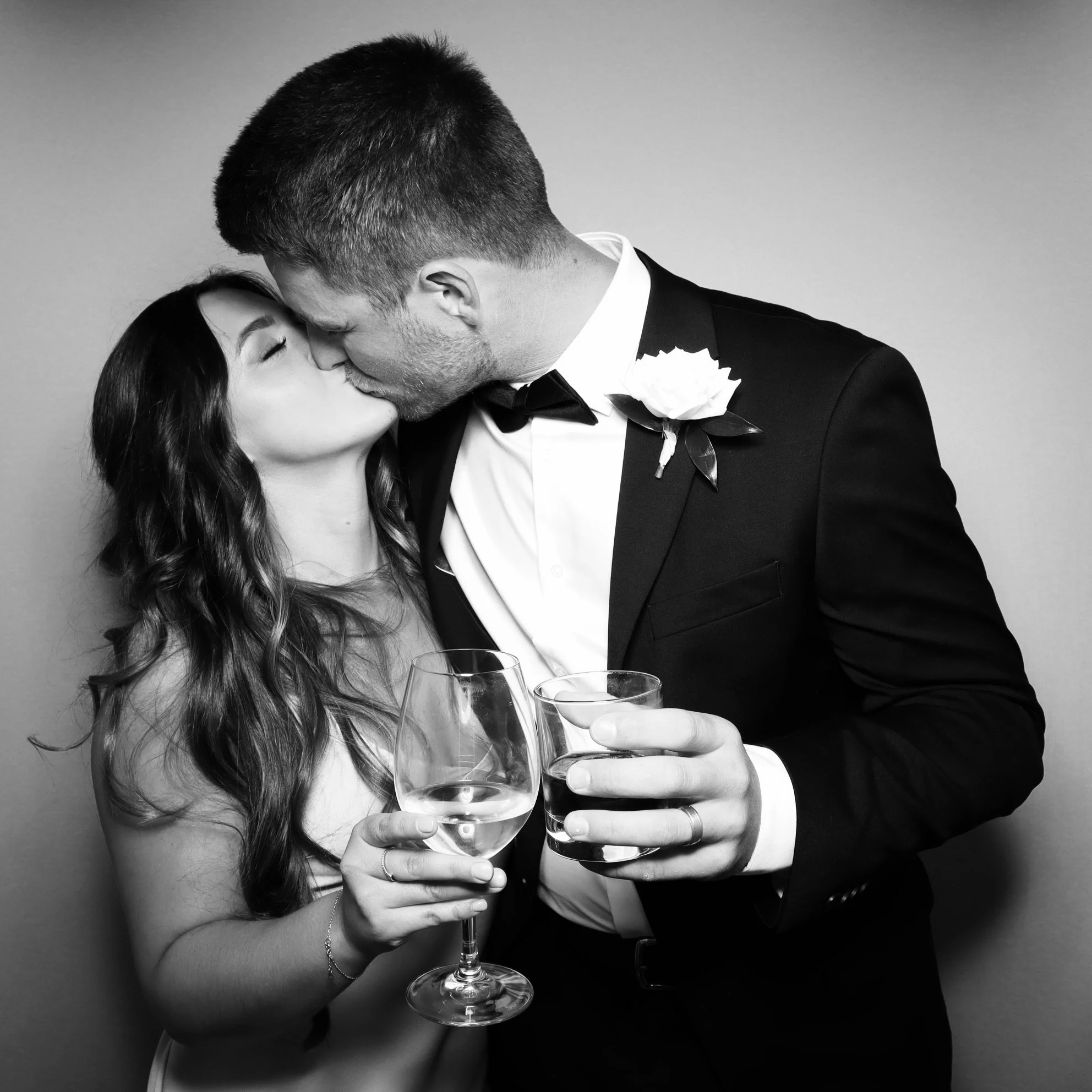 Black and white photo of a couple kissing at a wedding celebration, the woman holding a wine glass, the man dressed in a tuxedo with a boutonniere, both appear to be celebrating.