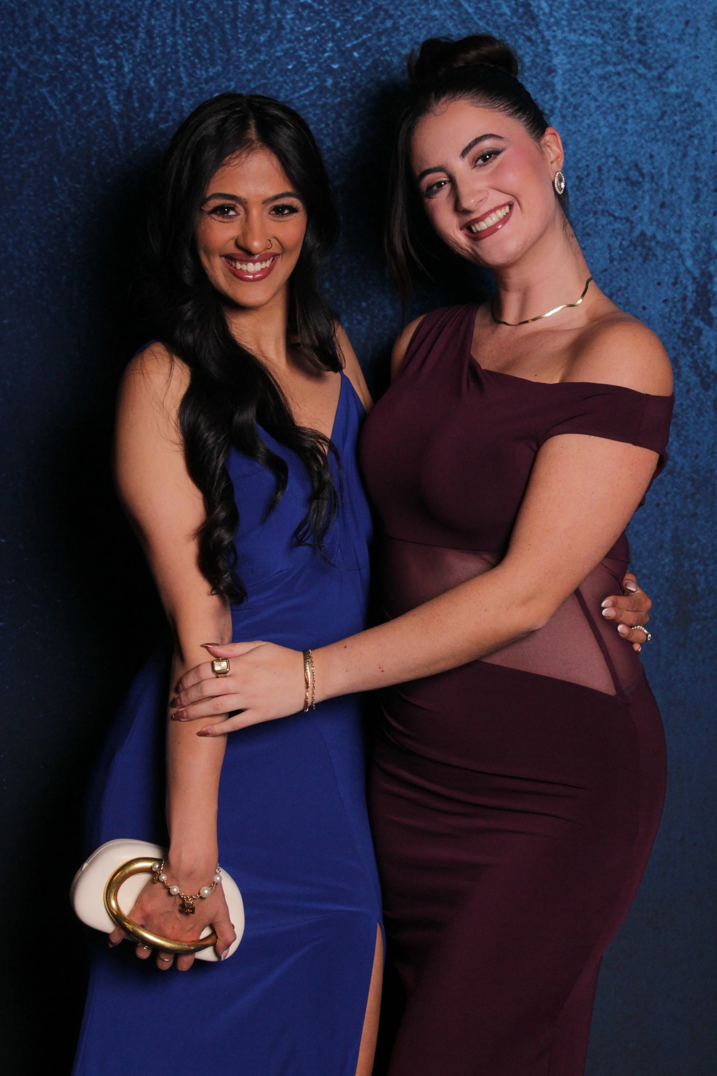 Two women in elegant dresses hugging against a blue textured background, smiling at the camera.