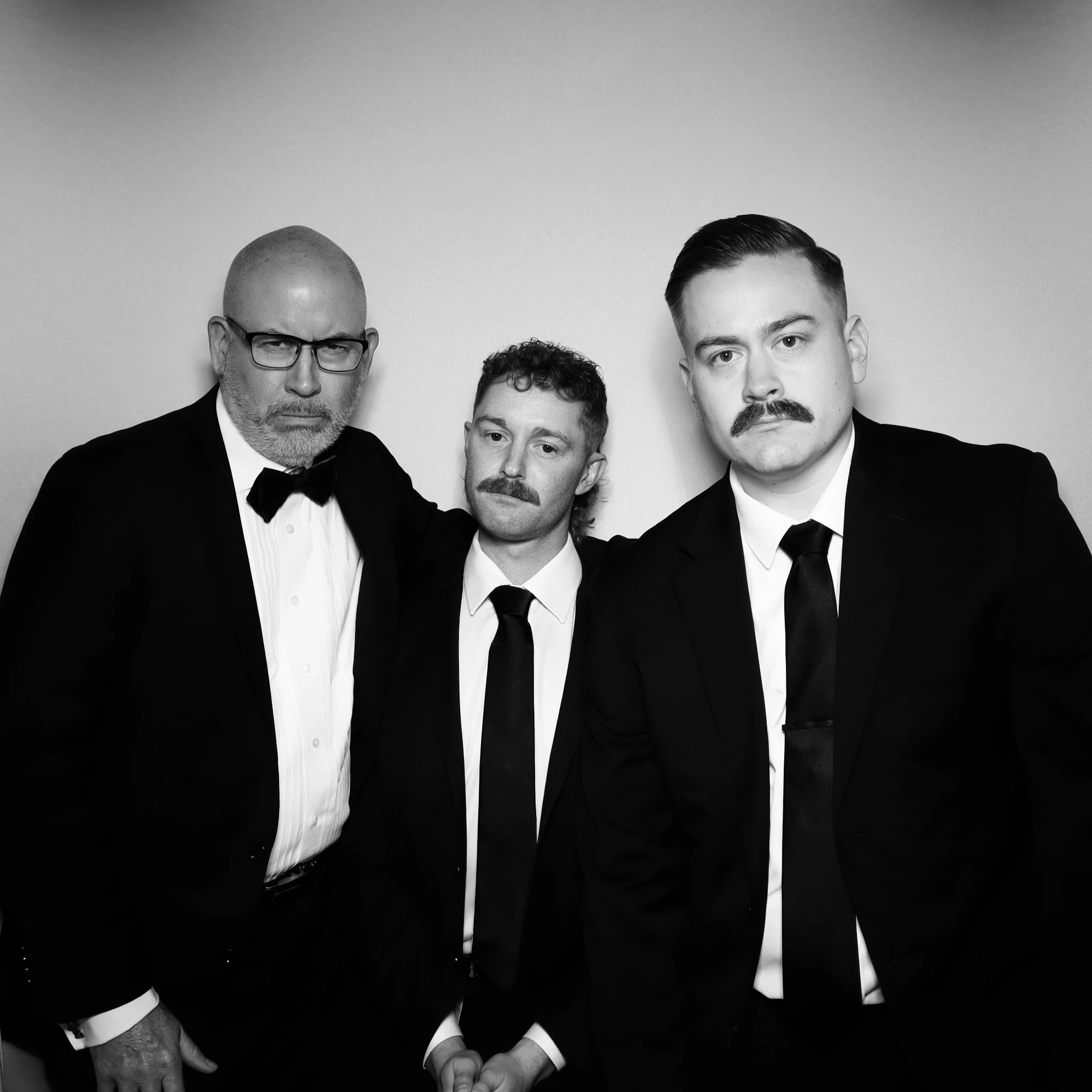 Three men in formal tuxedos with ties posing against a plain background.