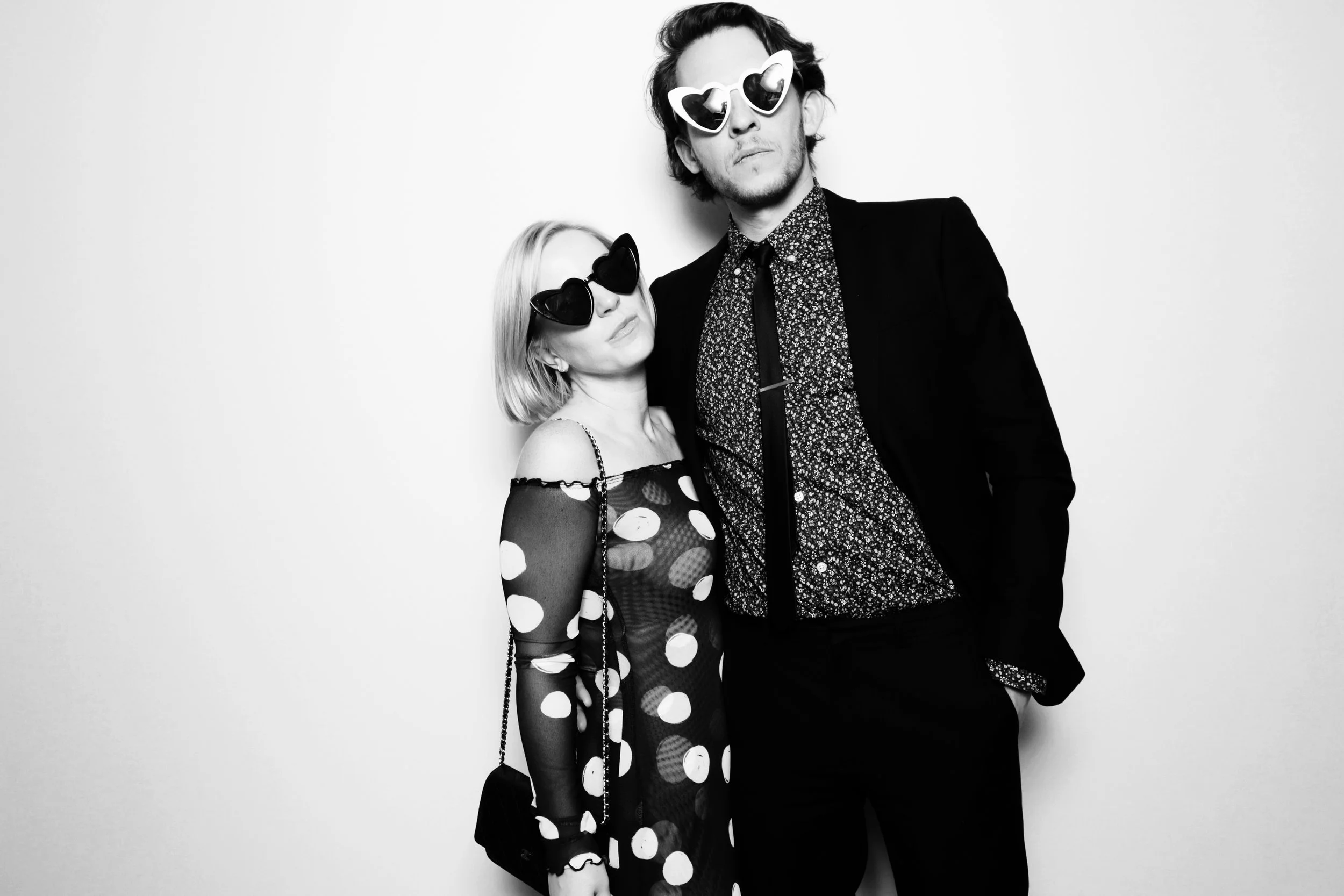 Black-and-white photo of a young woman and man wearing fashionable clothing and heart-shaped sunglasses, standing against a plain background.