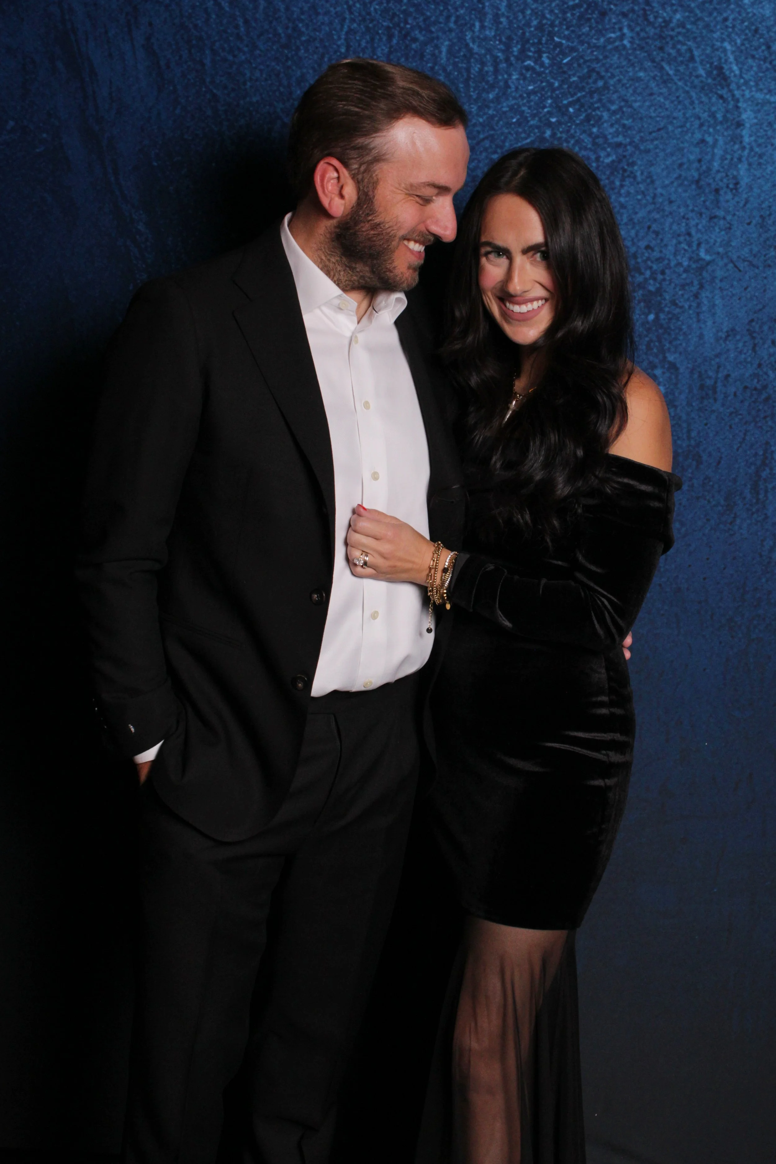 A man and woman dressed formally, smiling and embracing against a textured blue background.