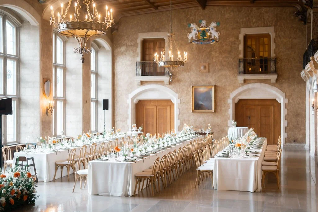 Elegant banquet hall with long tables, white tablecloths, floral centerpieces, and beige chairs, large chandeliers, stone walls, tall windows, and decorative coats of arms.