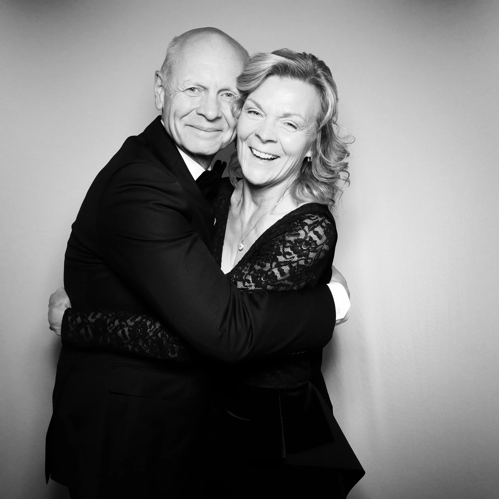 A black-and-white photograph of a happy, smiling older couple embracing each other, dressed in formal attire, against a plain background.