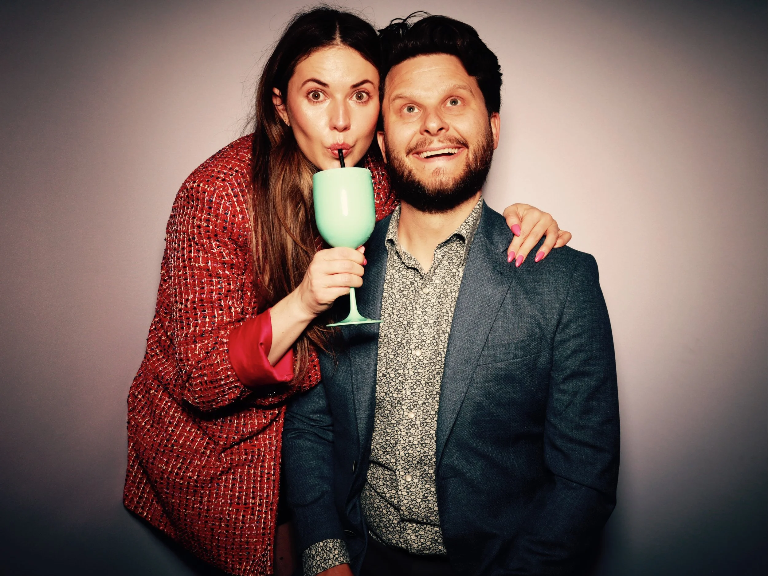 A woman with long brown hair and a woman with a short beard wearing a blue blazer and patterned shirt, smiling and holding a green drink with a straw, standing together against a neutral background.