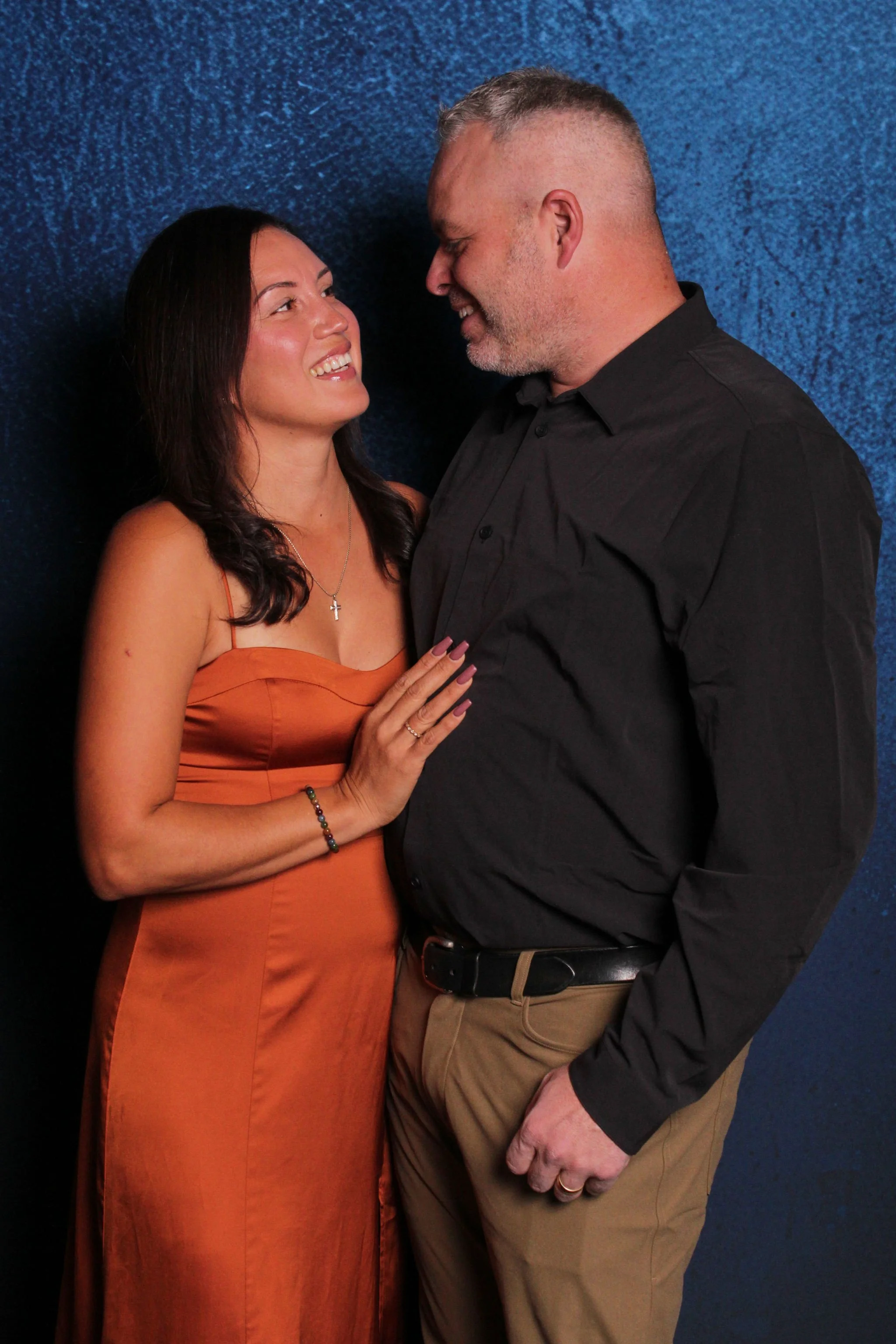 A couple standing close together, smiling and looking into each other's eyes against a dark blue textured background.