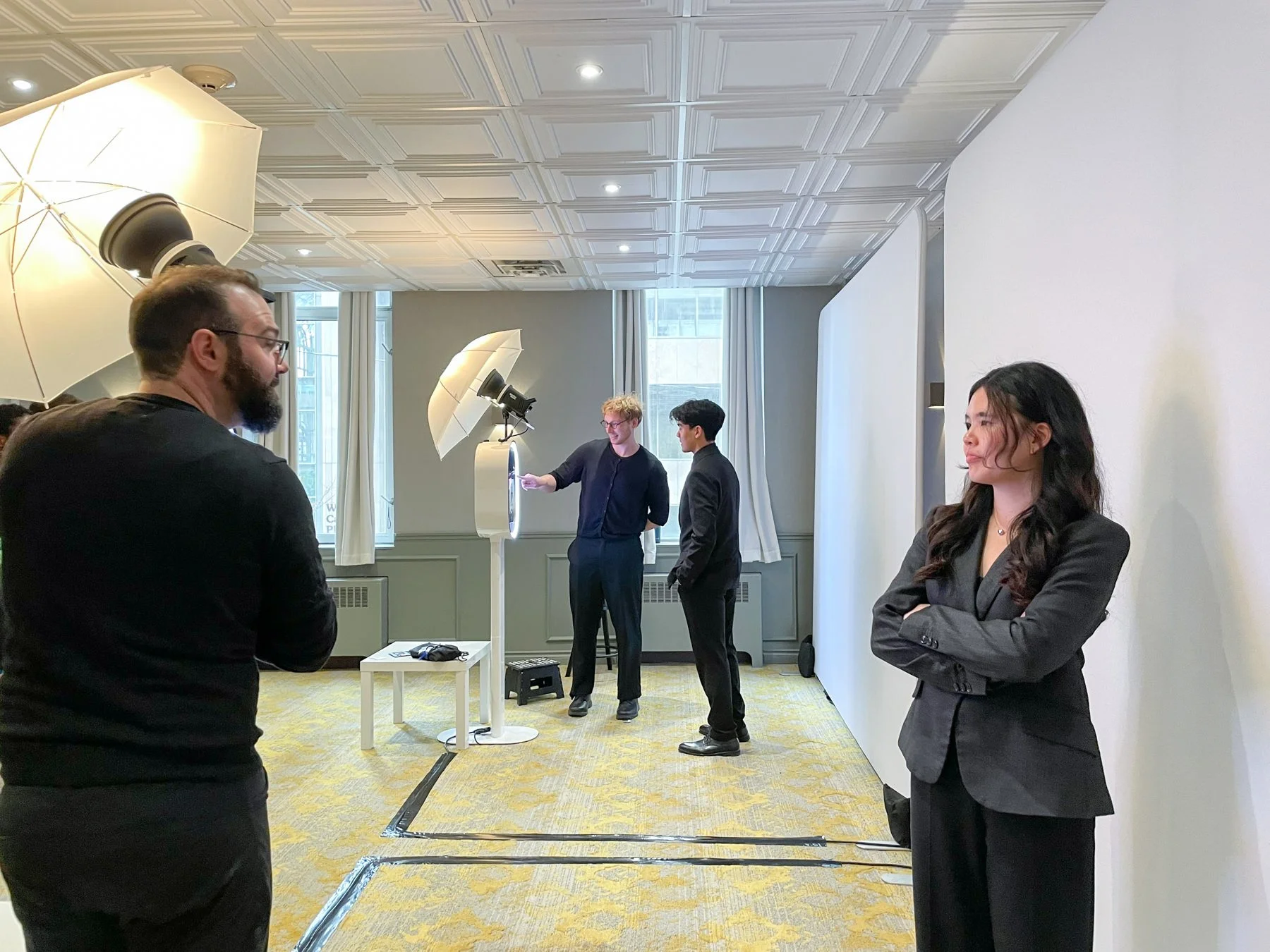 A professional photoshoot setup with a woman in a black blazer posing against a white backdrop, and two men in suits working with lighting equipment and a ring light, in a room with large windows and yellow patterned carpet.