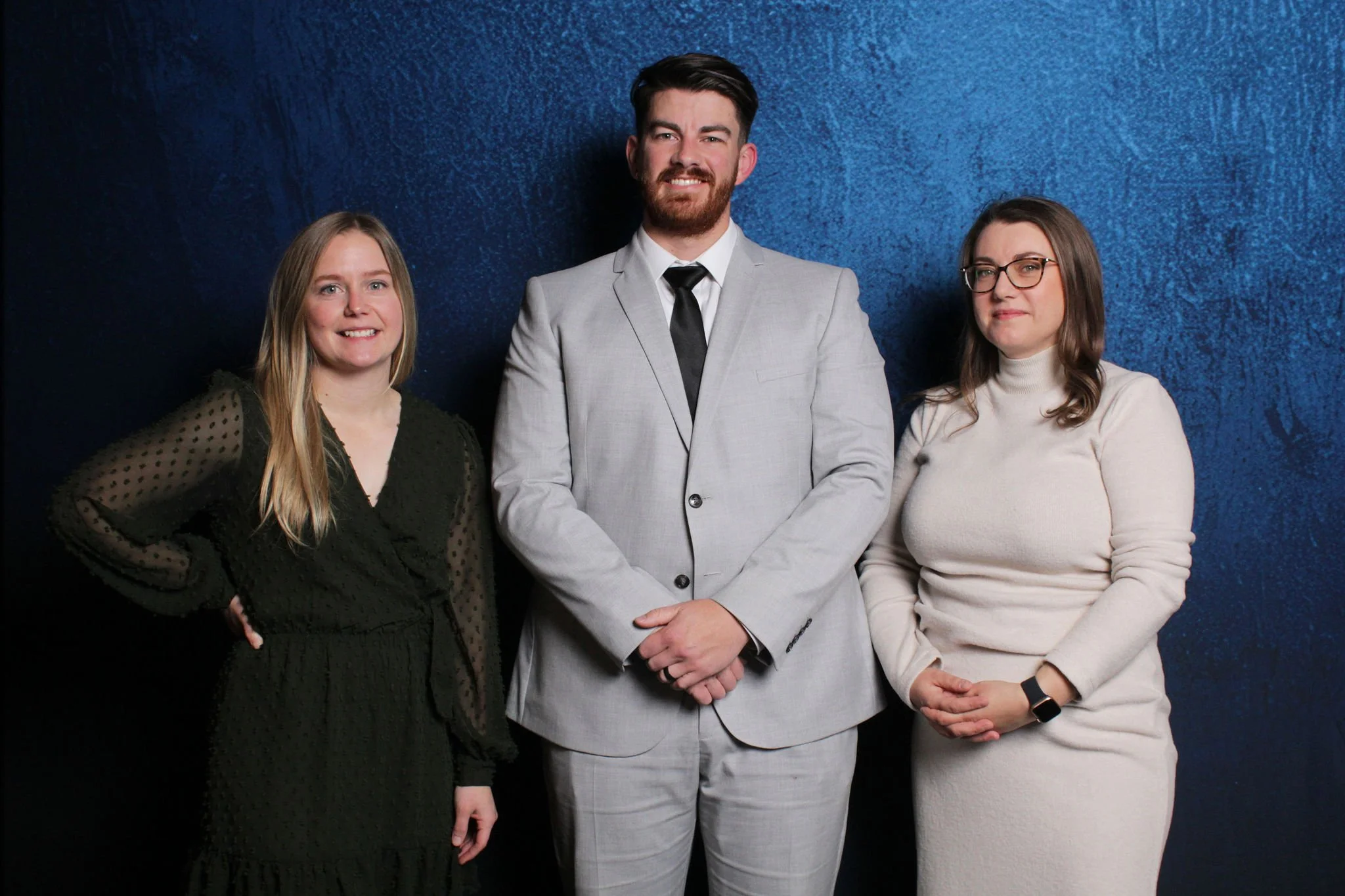 Three people standing against a textured blue background, a woman in a black dress on the left, a man in a light gray suit in the middle, and a woman in a white turtleneck sweater on the right.