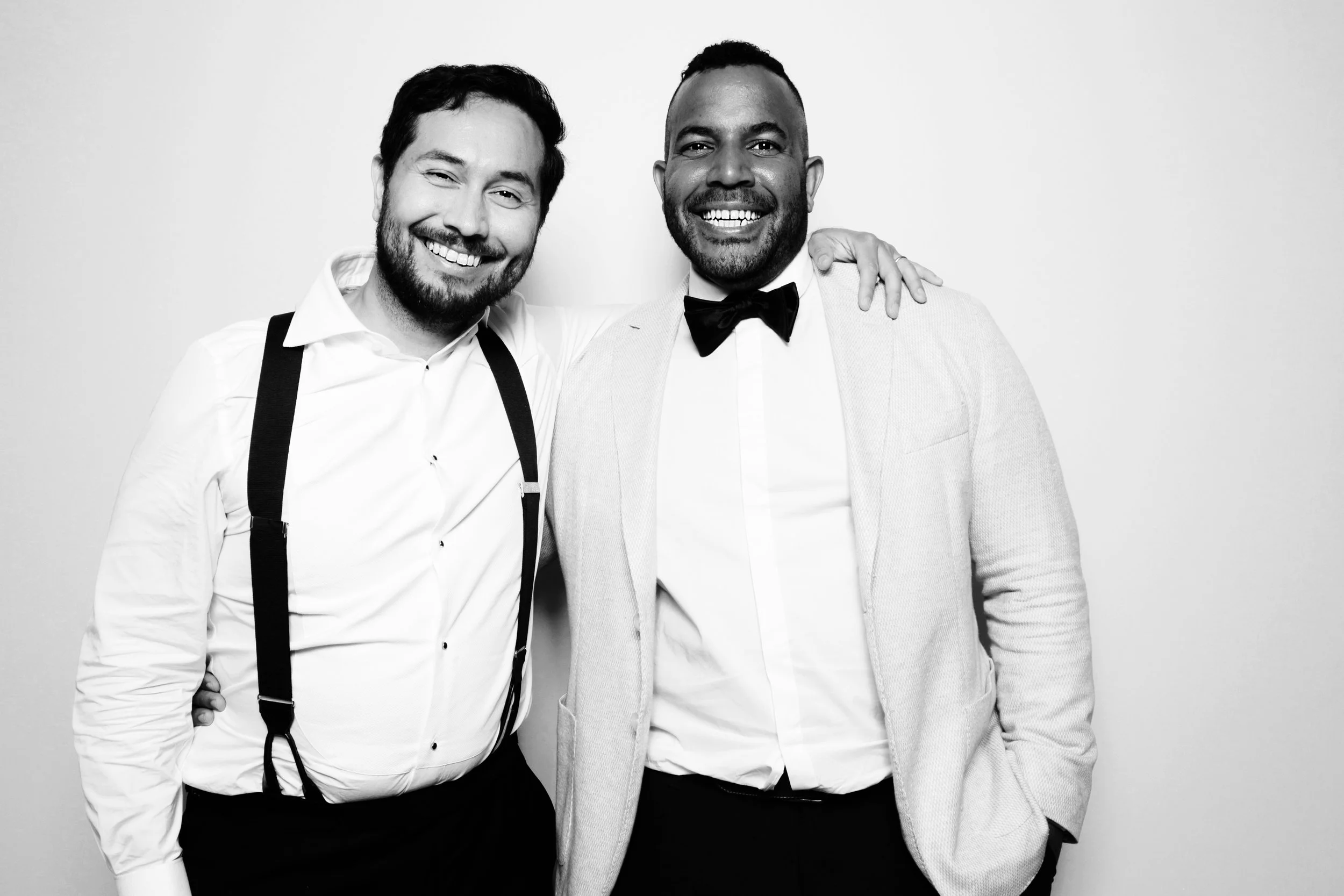 Two men smiling, one in a white shirt with suspenders and the other in a tuxedo jacket and bow tie, with their arms around each other in front of a plain background.