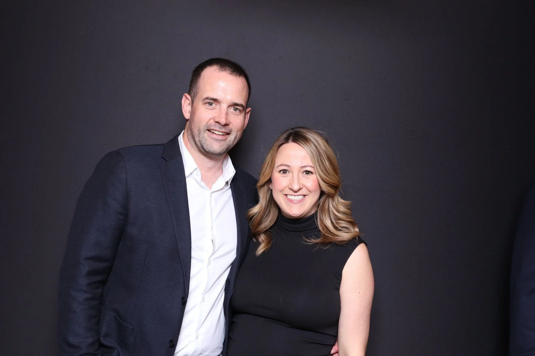 A man and woman smiling and standing together against a dark background.