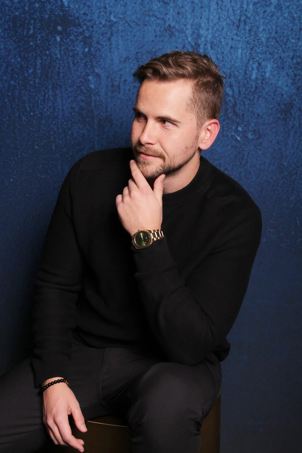 A man with short hair, a beard, and light skin is sitting against a textured blue background. He is wearing a black shirt, a gold watch, and a black beaded bracelet. He has his right hand on his chin and is looking to the left with a thoughtful expre
