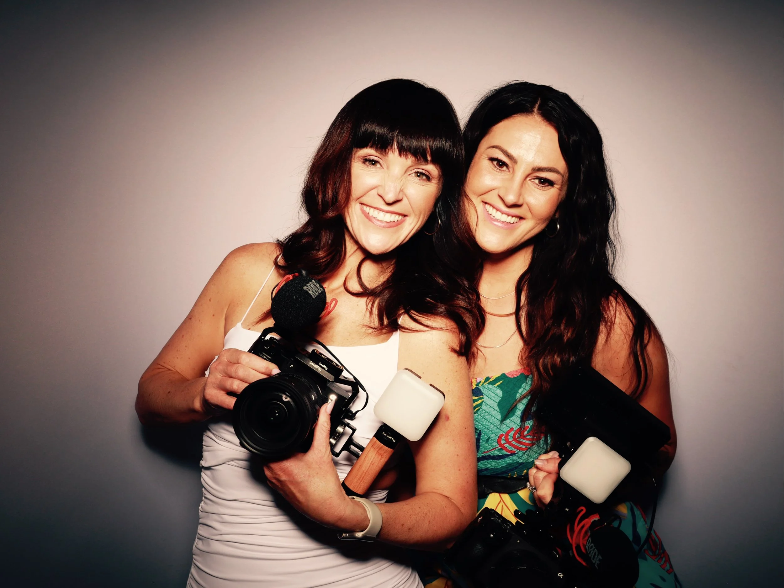 Two smiling women holding cameras and microphones, standing close together against a plain background.