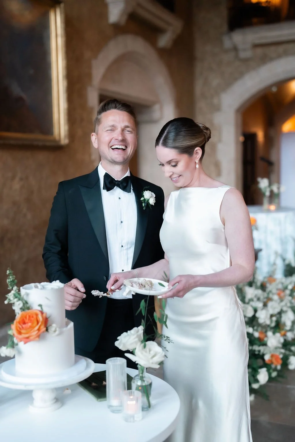 A newlywed couple cutting their wedding cake. The groom is wearing a black tuxedo with a bow tie, and the bride is in a white wedding dress. They are inside a decorated venue with flowers and warm lighting.