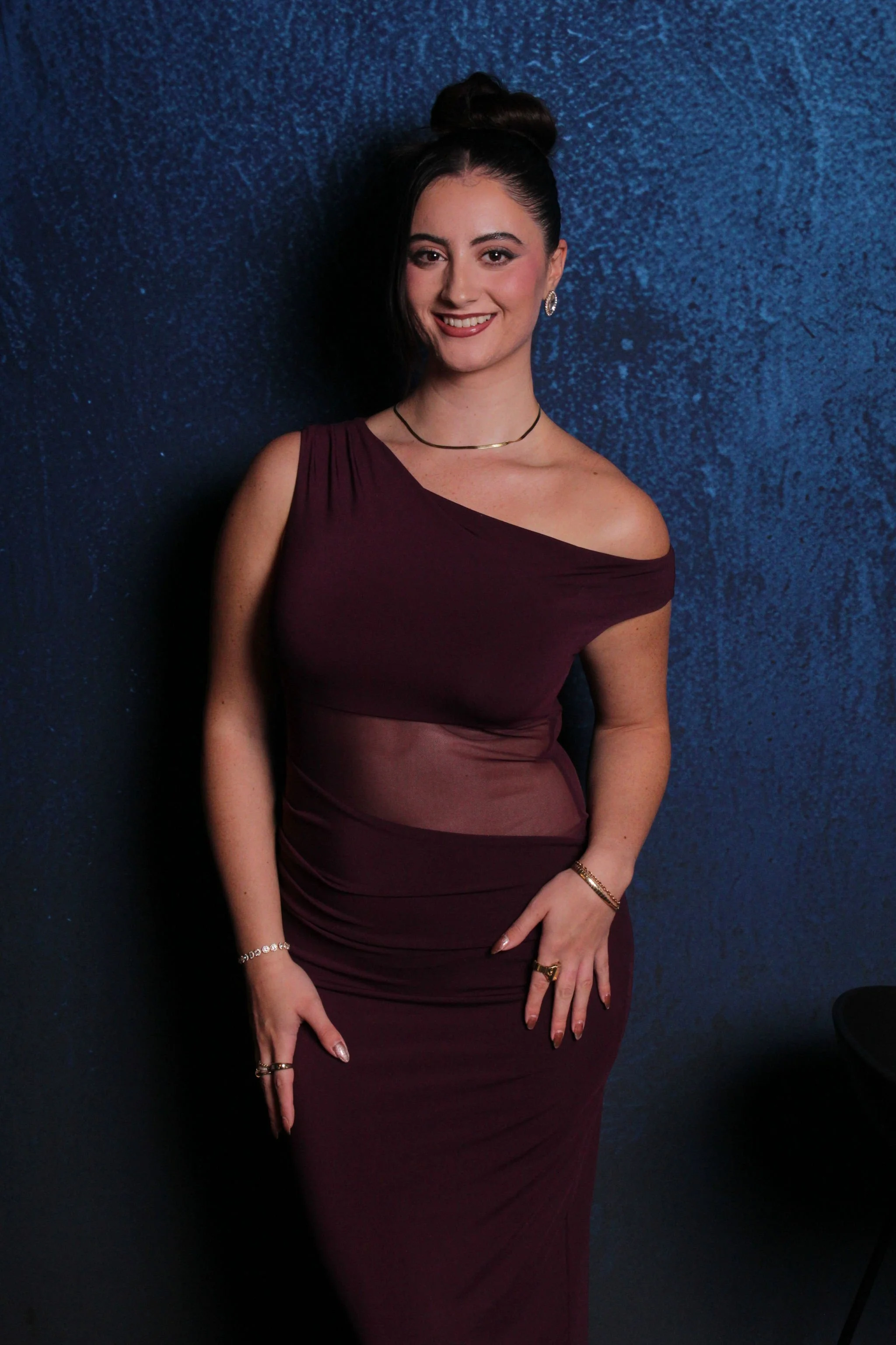 A woman in a burgundy dress standing against a dark blue textured wall, smiling at the camera.