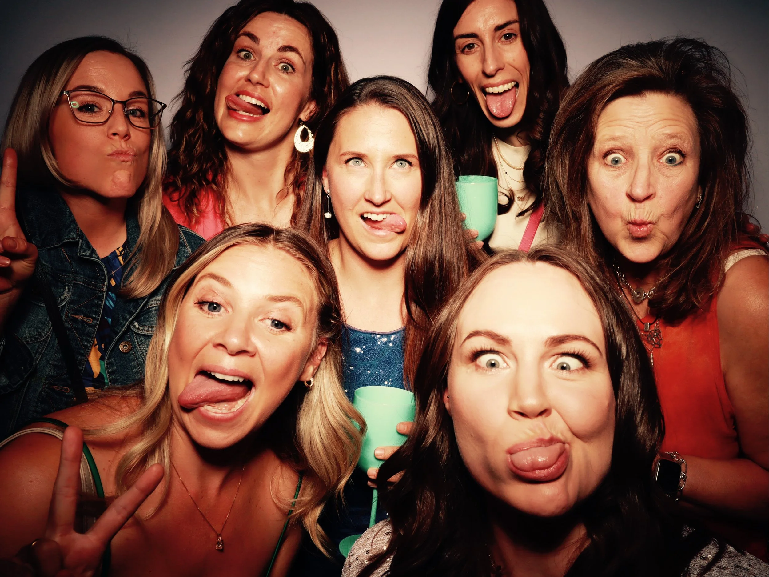 A group of eight women making silly faces, sticking out their tongues or puckering their lips, with some holding cups and making peace signs, against a plain background.