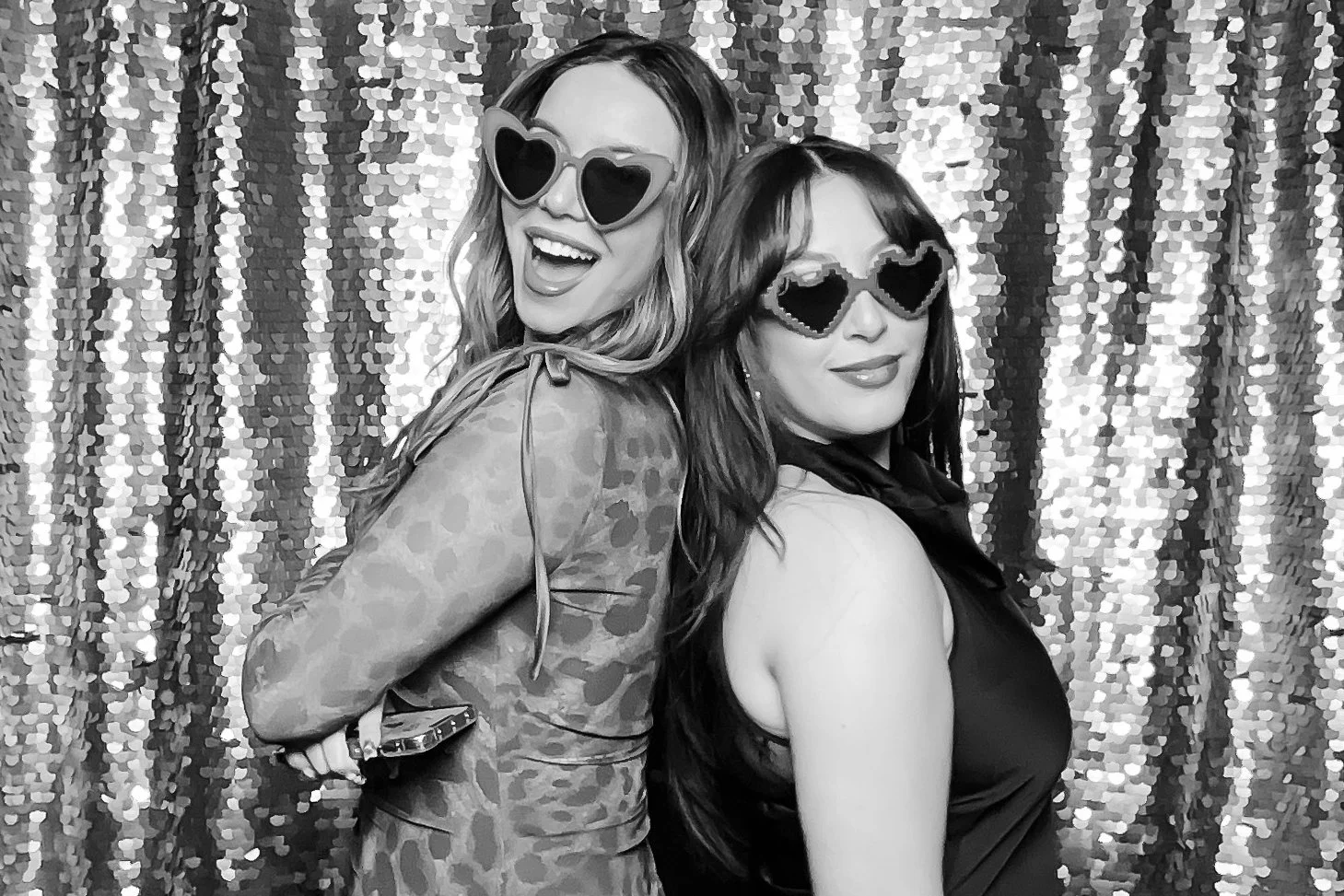 Two women posing with fun, heart-shaped sunglasses in front of a sequin backdrop, one smiling widely and the other with a subtle smile, both dressed stylishly for a lively event.