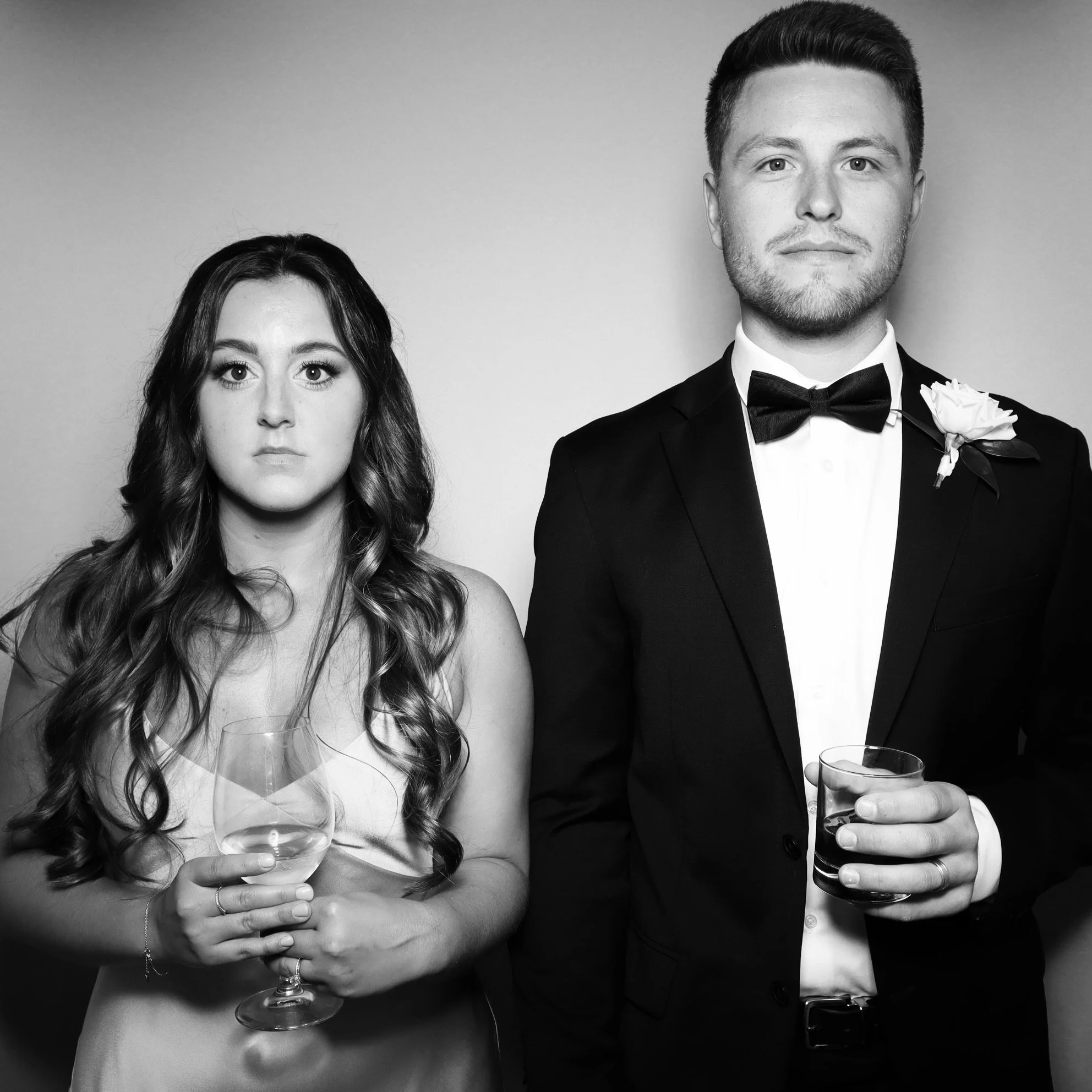 A black-and-white photo of a woman and a man dressed in formal attire holding drinks, standing against a plain background.