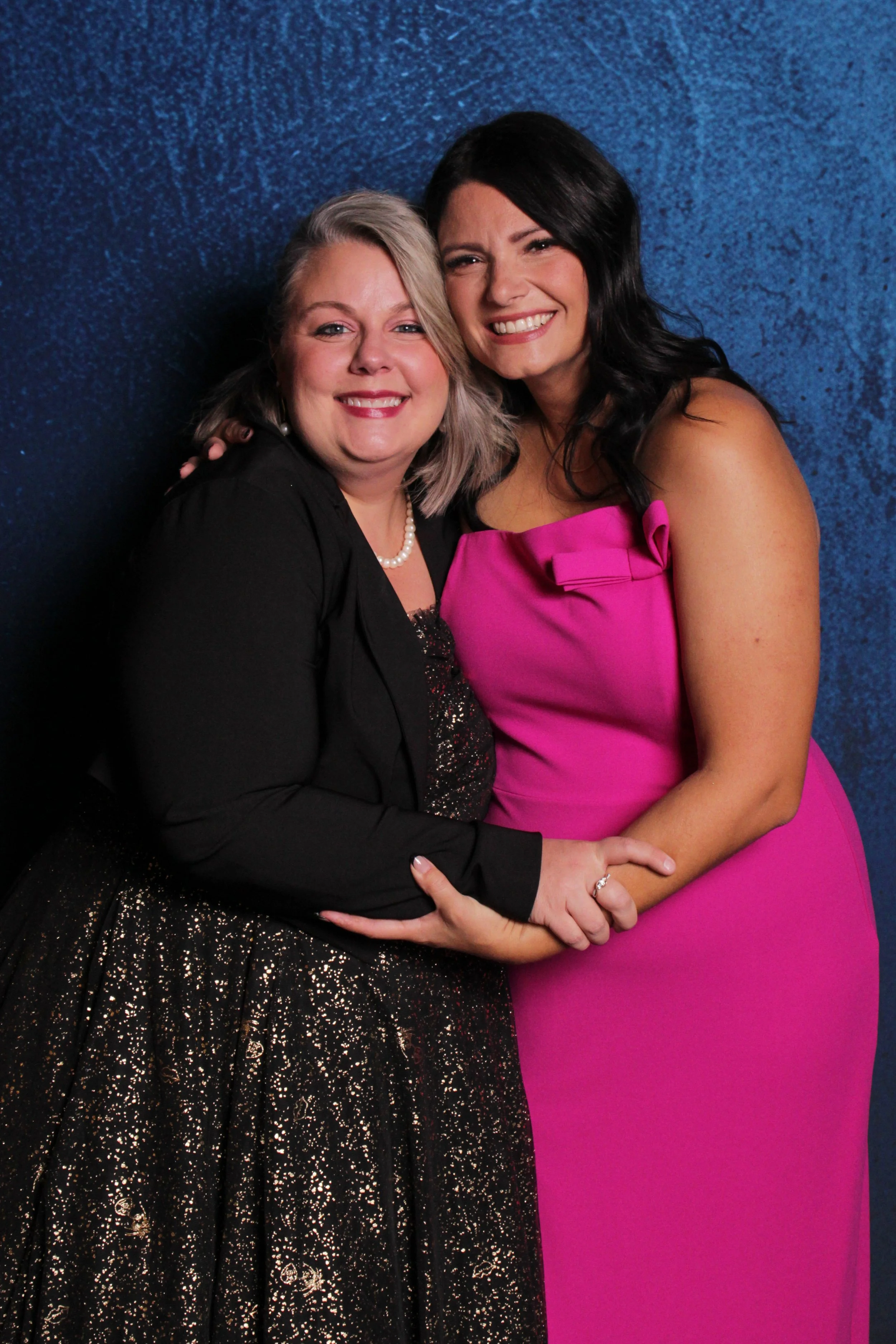 Two smiling women in formal attire standing close together against a dark blue textured background. The woman on the left has short, blonde hair, and is wearing a black dress with sequin details and a black blazer. She also has a pearl necklace. The 