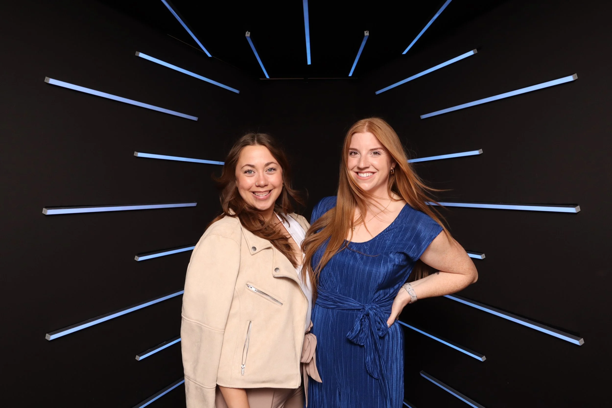 Two women standing together in front of a black wall with blue illuminated lines radiating outward, smiling at the camera.