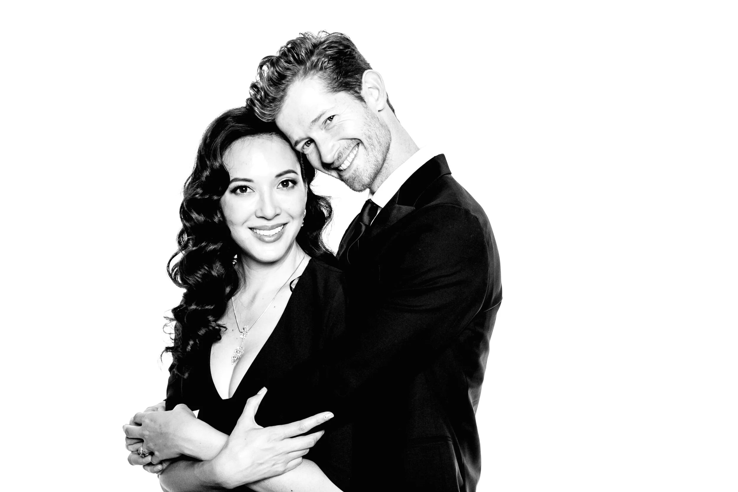 A black and white photo of a smiling woman with dark hair and a necklace, and a man with light hair and a beard, both dressed in formal attire, close together with the woman's arms crossed and the man embracing her.